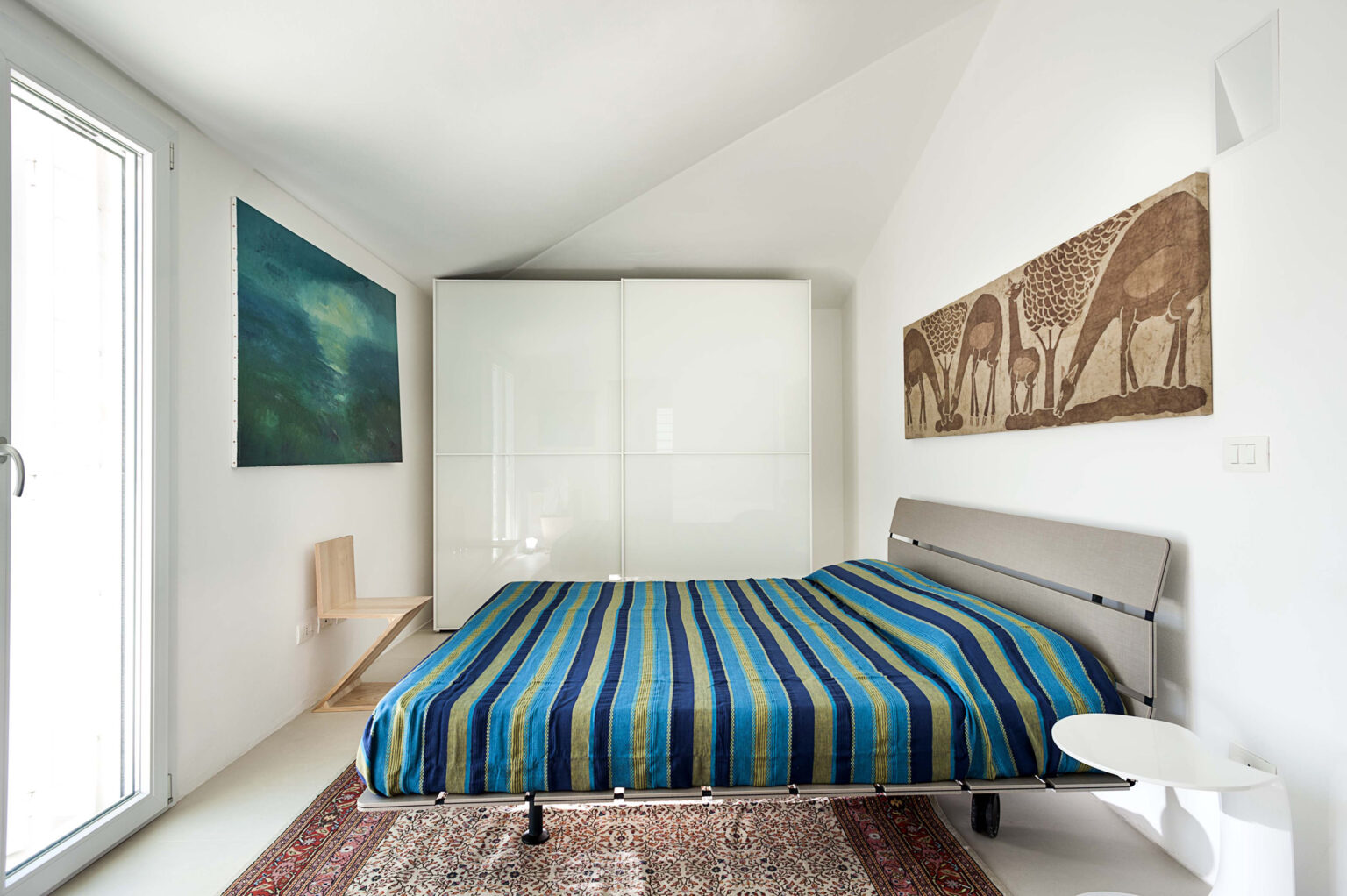 Modern bedroom featuring a striped blue and green bedspread, minimalist furniture, and artwork on the walls. The room includes large windows for natural light and a neutral color palette. | Only on SopranoVillas