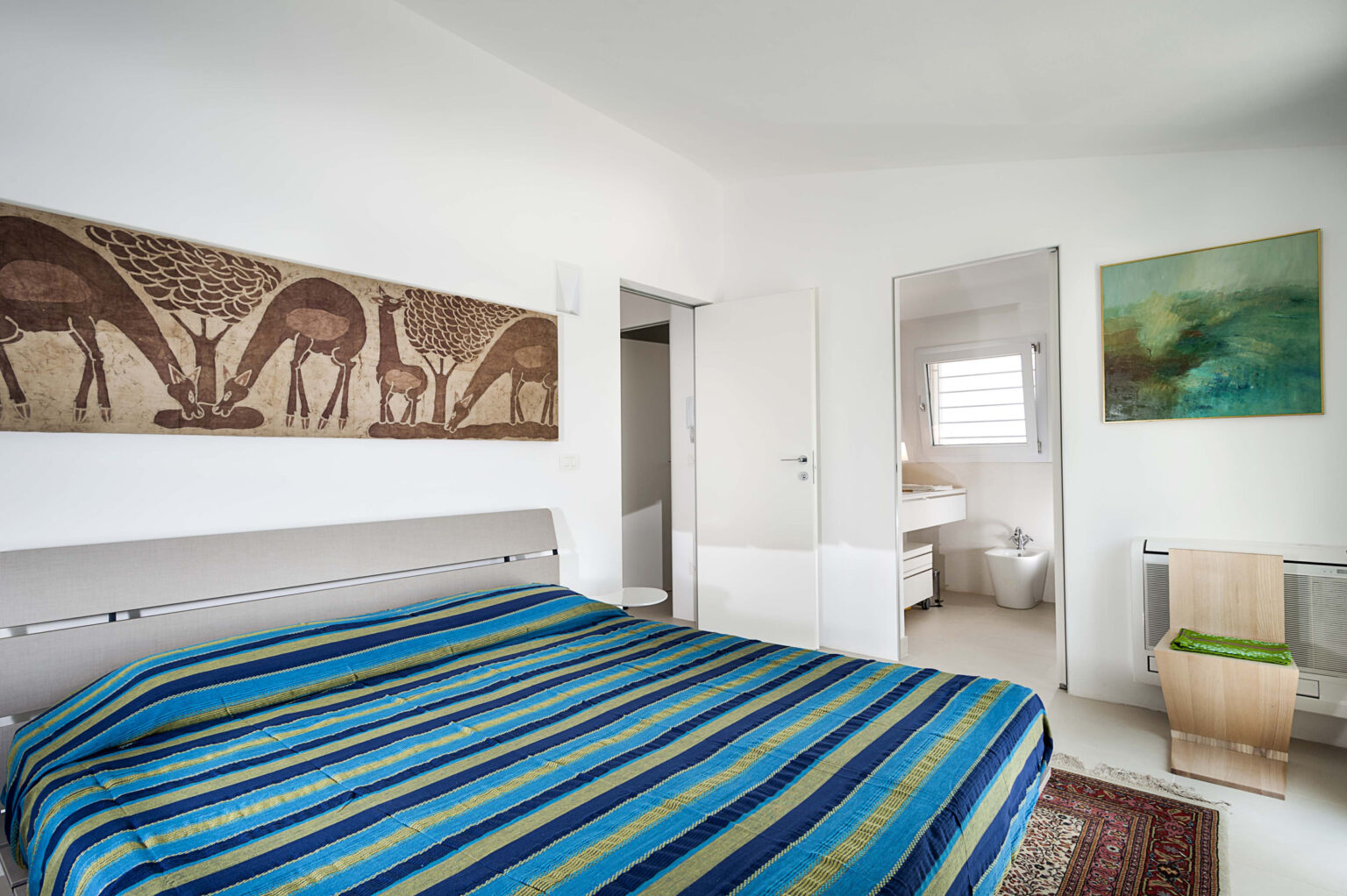 Modern bedroom featuring a striped blue and green bedspread, decorative wall art of giraffes, and minimalist furniture. The room includes an en-suite bathroom and a neutral color scheme, emphasizing a spacious and inviting atmosphere. | Only on SopranoVillas