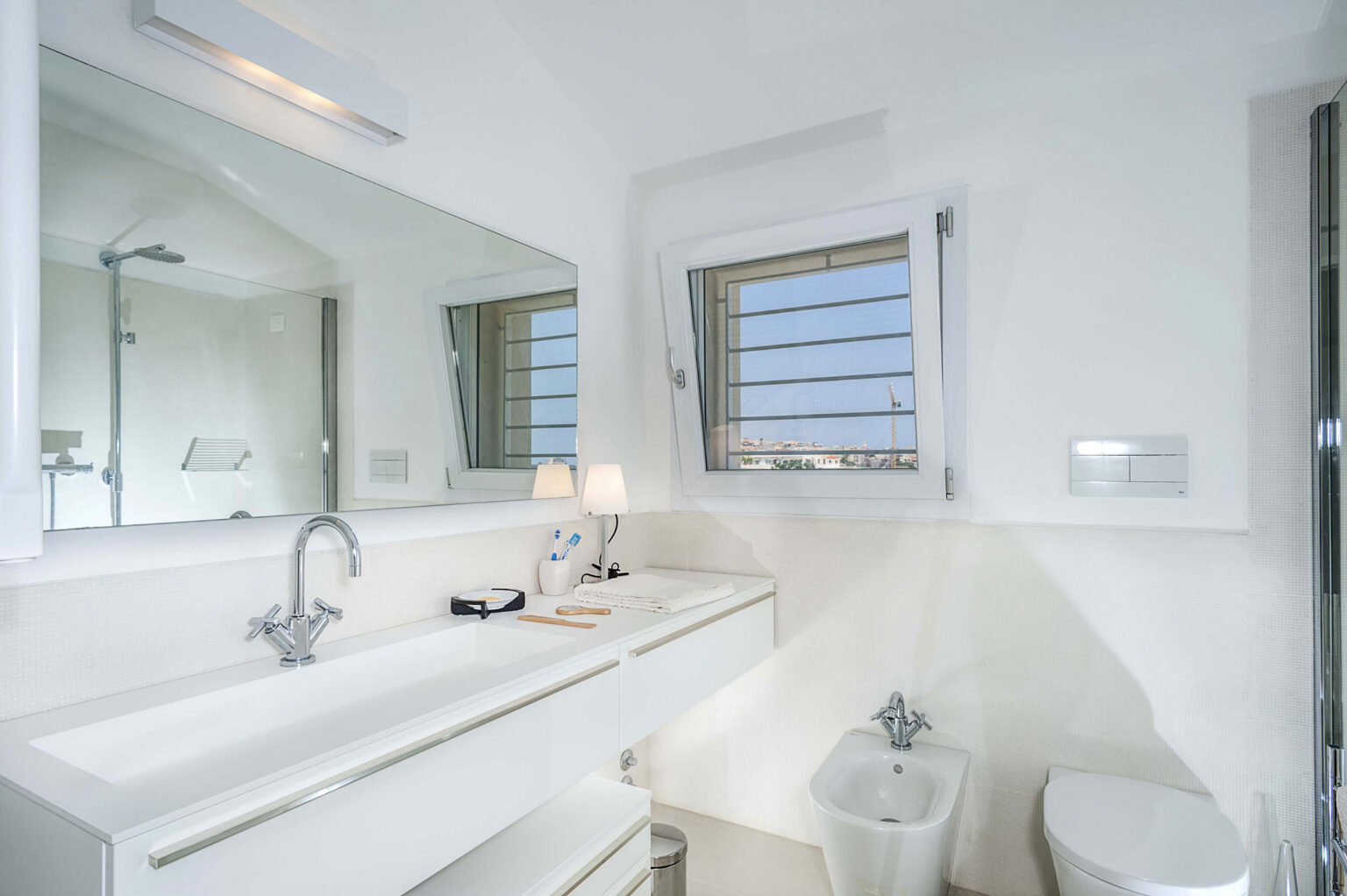 Modern bathroom featuring a white vanity with a sink, a large mirror, a shower area, and a window with shutters, allowing natural light to illuminate the space. | Only on SopranoVillas