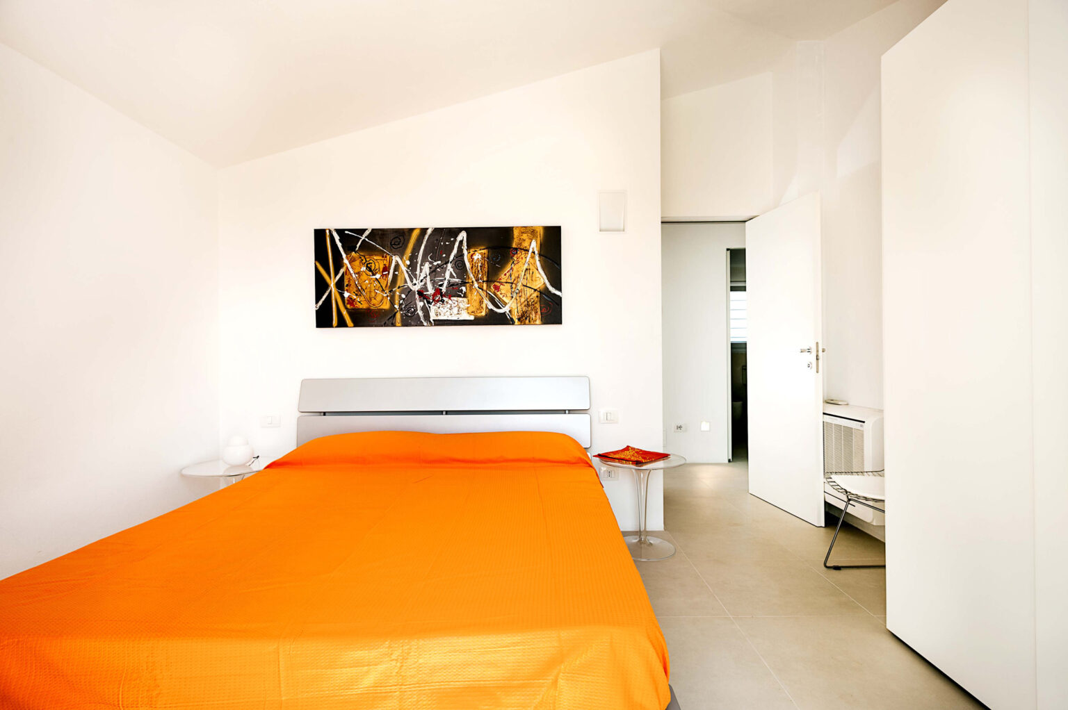 Modern bedroom featuring an orange bedspread, minimalist decor, and abstract wall art. The room has white walls, a small side table, and a doorway leading to a bathroom, showcasing a sleek and contemporary design. | Only on SopranoVillas