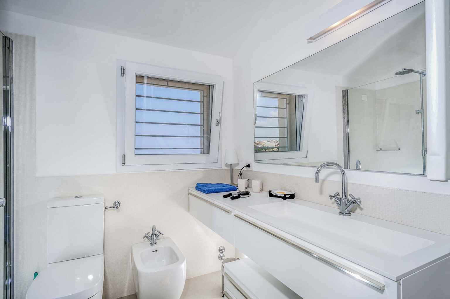 Modern bathroom featuring a sleek design with a white vanity, large mirror, and ample natural light from windows. The space includes a toilet, bidet, and glass shower enclosure, complemented by blue towels and toiletries on the countertop. | Only on SopranoVillas