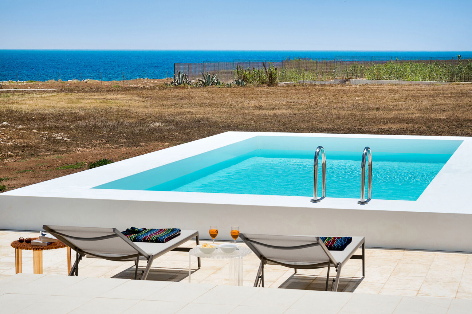 Modern outdoor pool overlooking the ocean, featuring two lounge chairs and a scenic landscape. Ideal for relaxation and enjoying coastal views. | Only on SopranoVillas