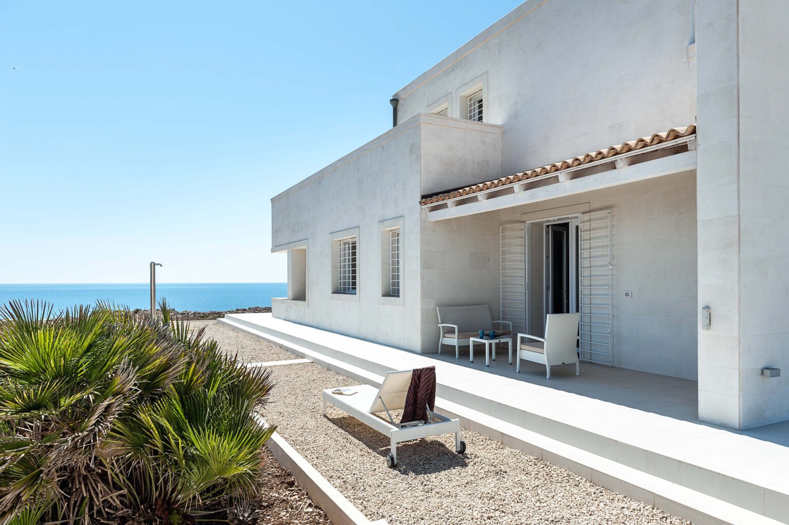 Modern seaside villa with a spacious terrace, featuring outdoor seating and a sun lounger, surrounded by gravel landscaping and palm plants, offering stunning ocean views under a clear blue sky. | Only on SopranoVillas