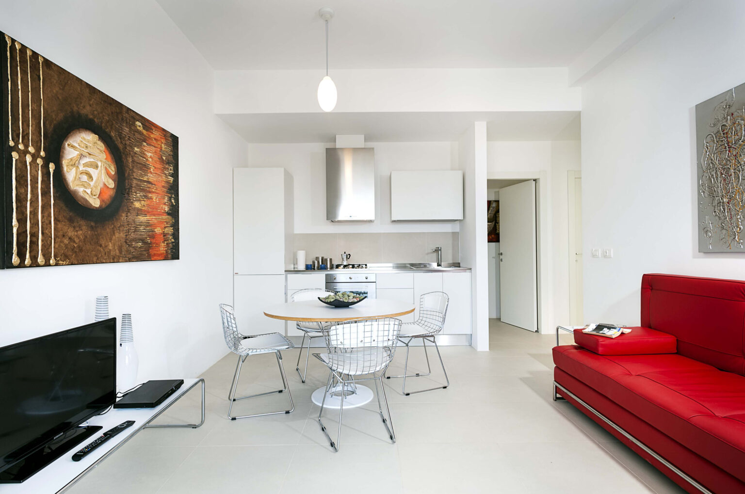 Modern living room and kitchen area featuring a red sofa, a round dining table with wire chairs, and contemporary artwork on the walls. The kitchen is equipped with stainless steel appliances and white cabinetry, creating a bright and inviting space. | Only on SopranoVillas