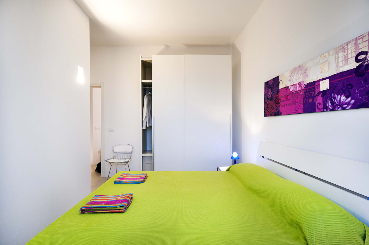 Modern bedroom featuring a vibrant green bedspread, colorful towels, and a stylish white wardrobe. The room includes a small chair and a decorative wall art piece, creating a bright and inviting atmosphere. Ideal for relaxation and comfort. | Only on SopranoVillas