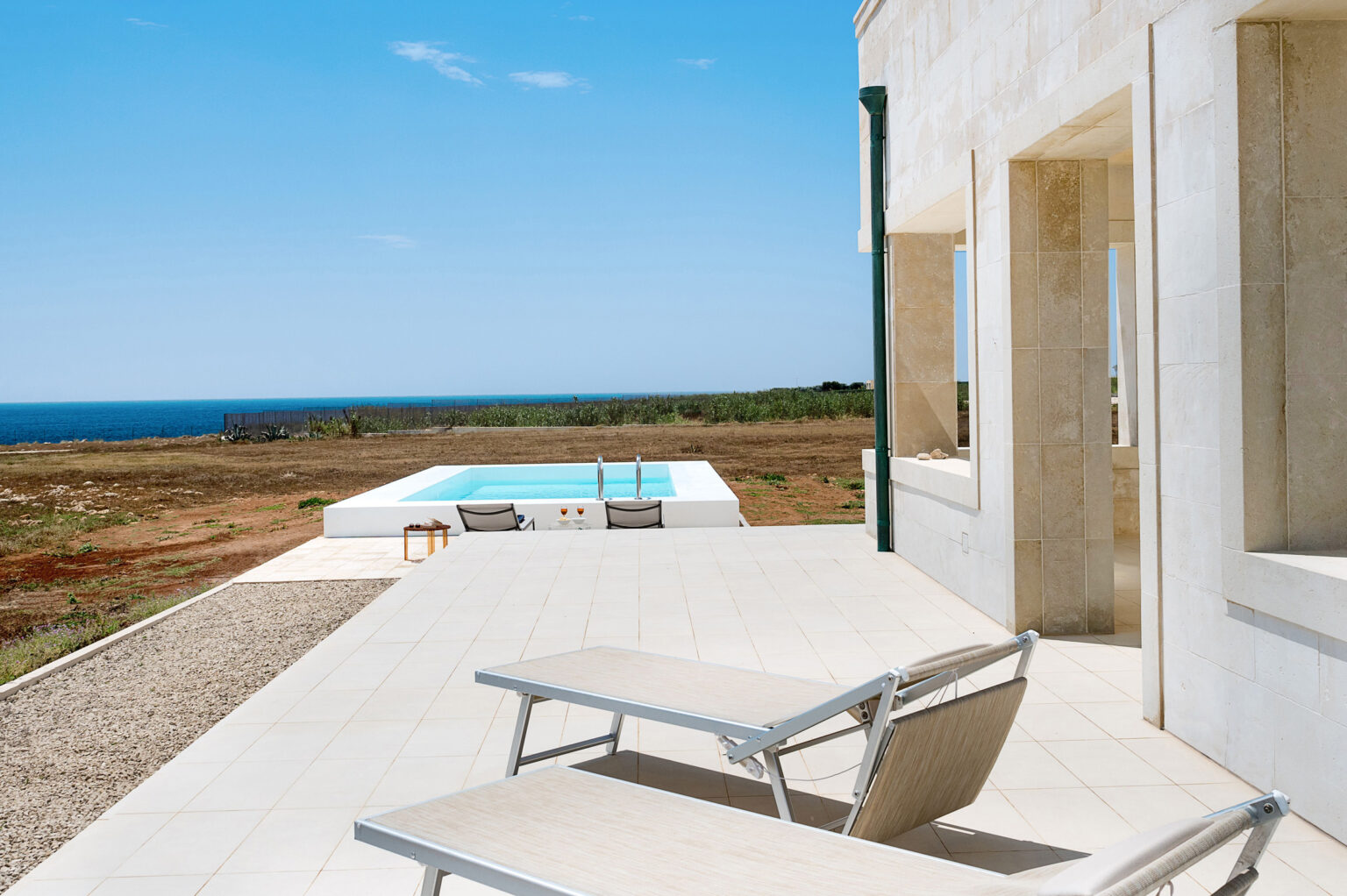 Modern vacation home terrace with lounge chairs overlooking a serene pool and ocean view, surrounded by lush landscape under a clear blue sky. | Only on SopranoVillas