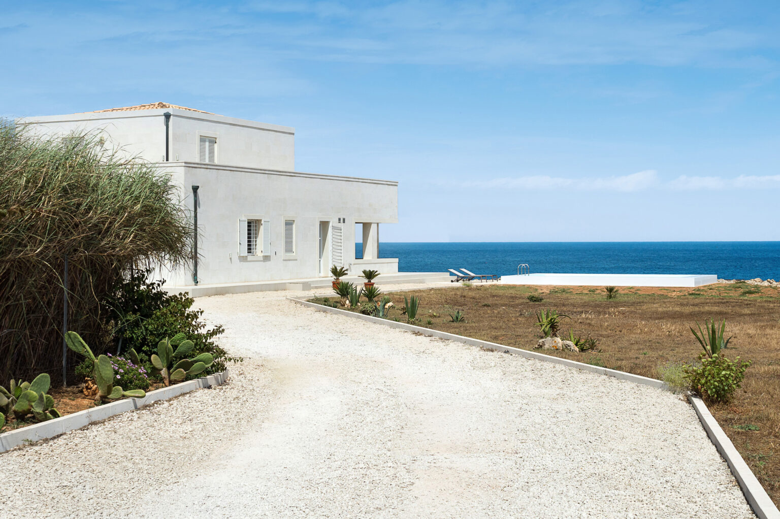 Modern coastal villa with a gravel pathway leading to a sea view, featuring a swimming pool and landscaped garden with cacti and greenery. Ideal for a serene getaway by the ocean. | Only on SopranoVillas