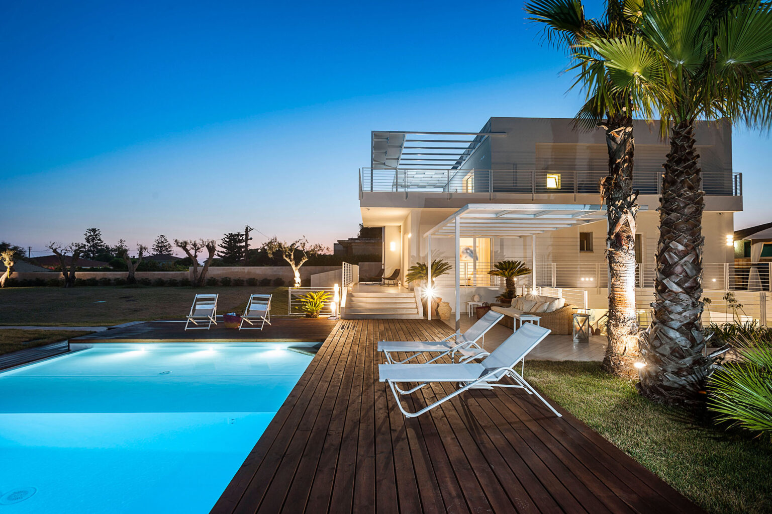 Modern luxury villa with a swimming pool at sunset, featuring palm trees and lounge chairs on a wooden deck, ideal for relaxation and outdoor living. | Only on SopranoVillas