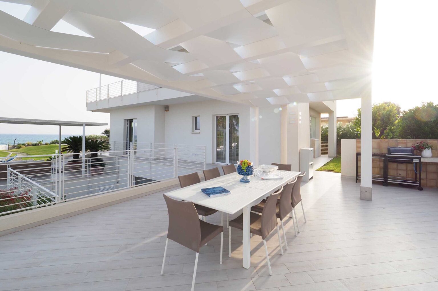Modern outdoor dining area with a white table and beige chairs, featuring a blue vase with flowers, under a stylish canopy. The setting offers a view of the ocean and lush greenery, providing an inviting atmosphere for alfresco meals. | Only on SopranoVillas
