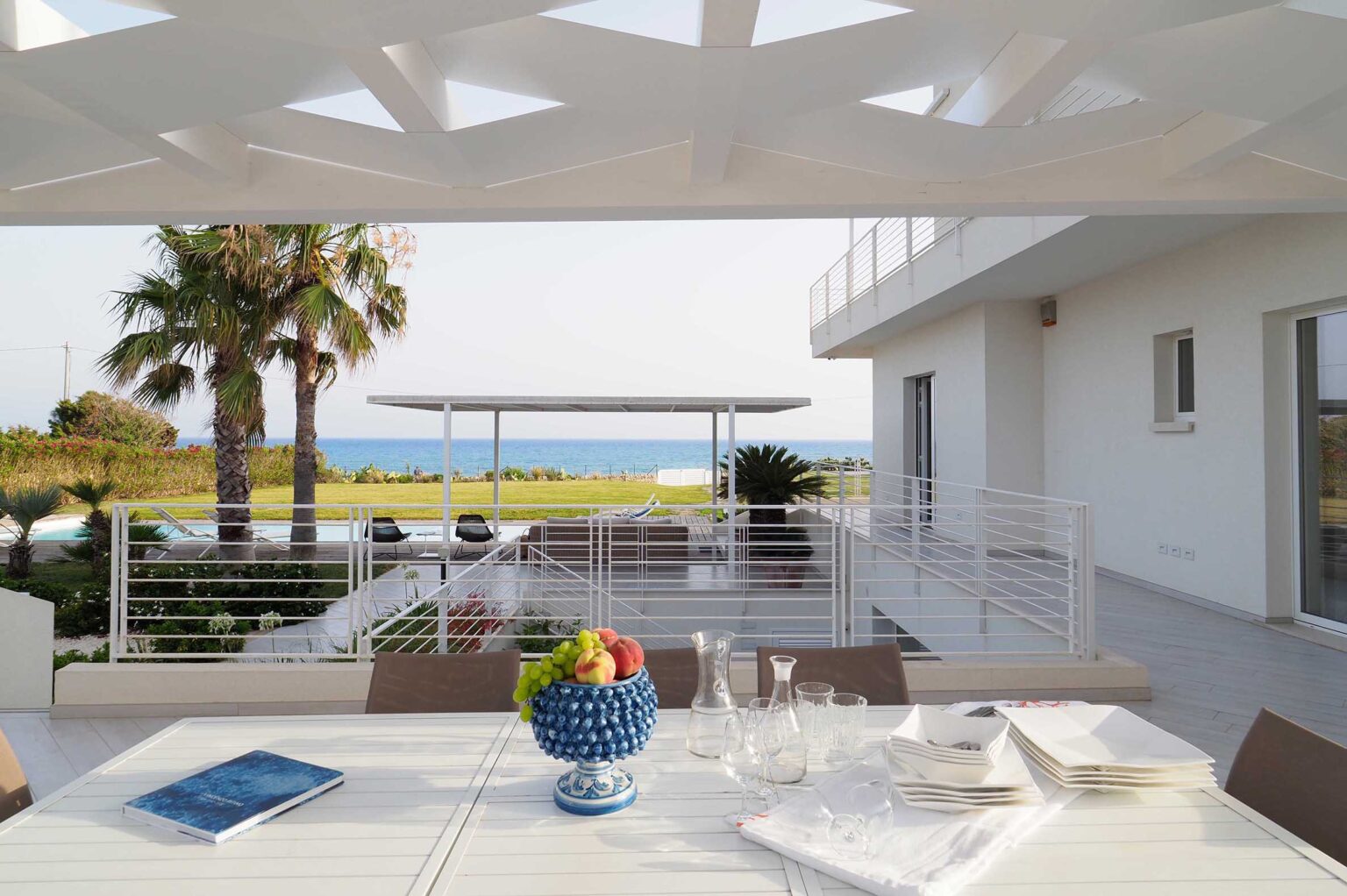 Modern outdoor dining area with a view of the ocean, featuring a stylish white table set with a blue fruit bowl, glassware, and a book. Lush palm trees and landscaped gardens enhance the serene atmosphere, creating an inviting space for relaxation. | Only on SopranoVillas