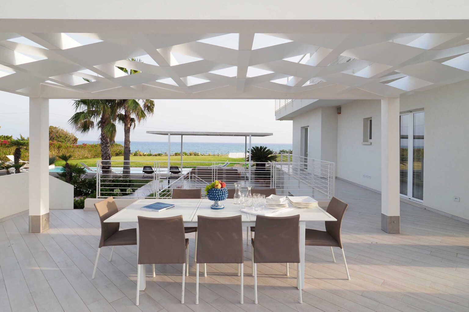 Modern outdoor dining area with a white table set for six, surrounded by gray chairs, overlooking a scenic view of the ocean and palm trees. The pergola features a geometric design, allowing natural light to filter through. Ideal for alfresco dining and entertaining. | Only on SopranoVillas