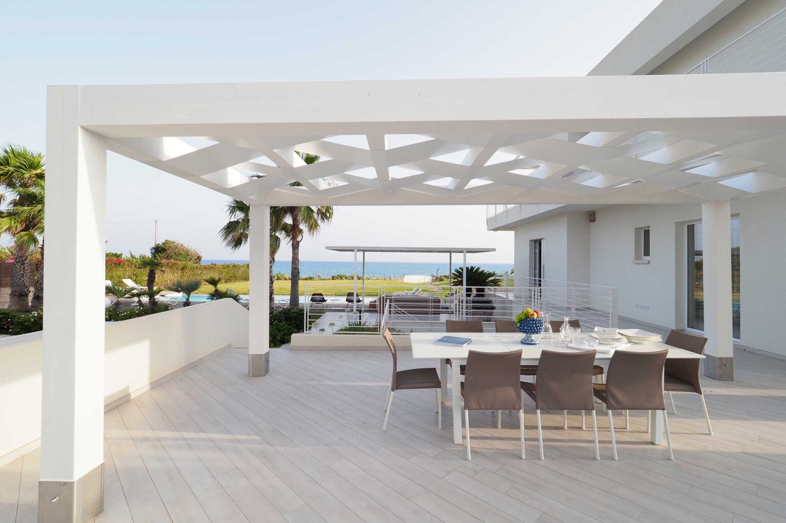 Modern outdoor dining area with a white pergola, featuring a table set for a meal and surrounded by palm trees, overlooking a scenic view of the ocean and landscaped garden. Perfect for entertaining and enjoying coastal living. | Only on SopranoVillas
