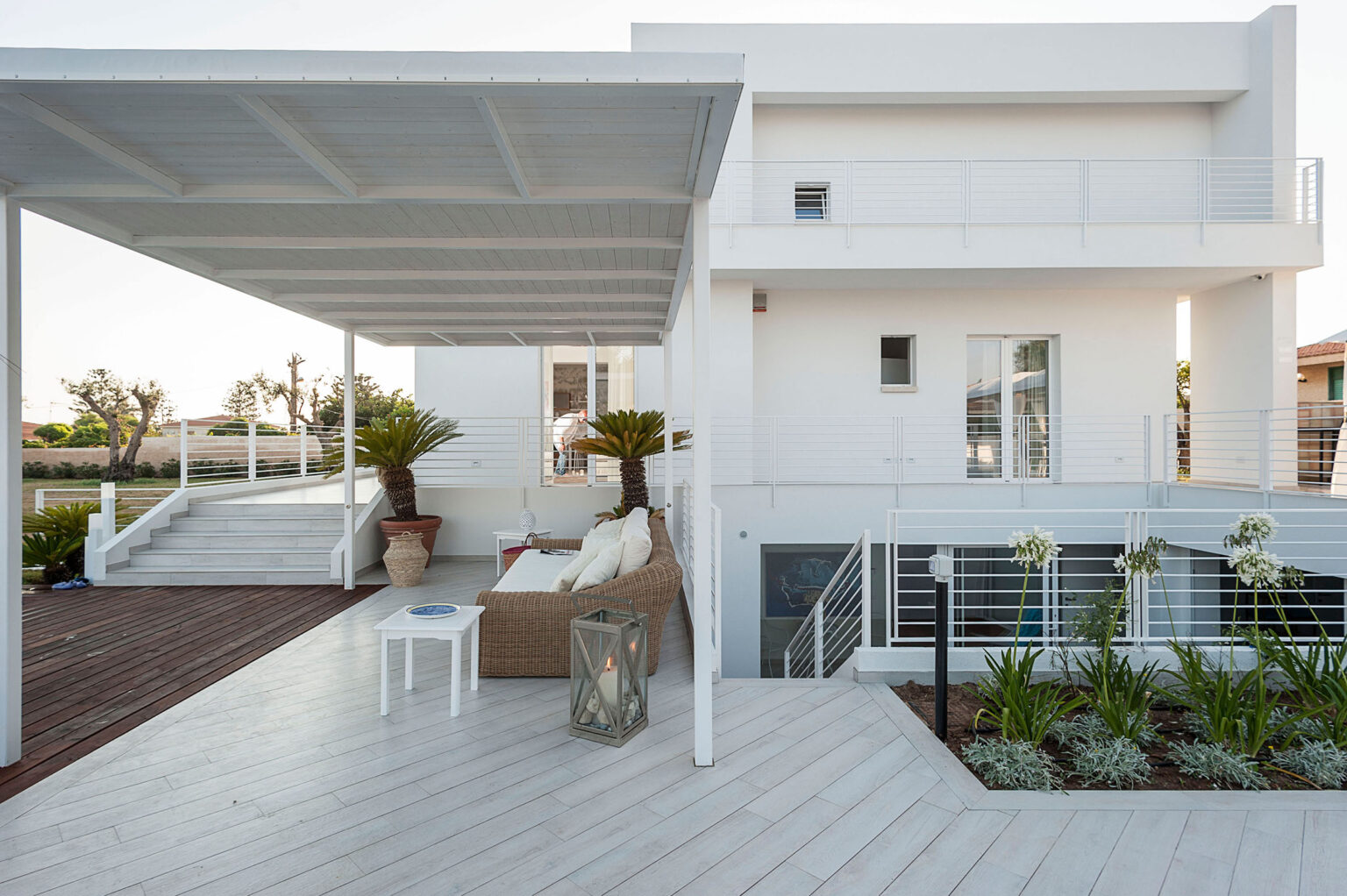 Modern outdoor living space featuring a white house with a spacious patio, comfortable seating, and lush greenery. The design emphasizes clean lines and an inviting atmosphere, perfect for relaxation and entertaining. | Only on SopranoVillas