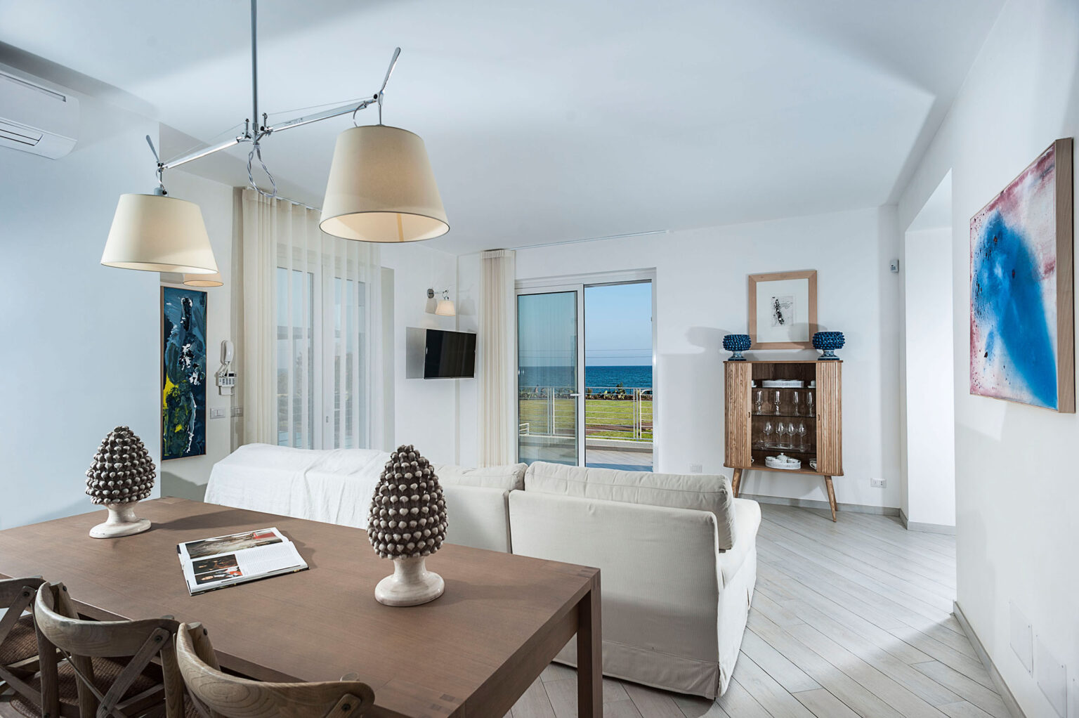 Bright and airy living room featuring modern decor, a dining table with decorative pine cone centerpieces, and large windows offering a view of the ocean. The space includes a comfortable sofa, wall art, and a television, creating a welcoming atmosphere for relaxation. | Only on SopranoVillas