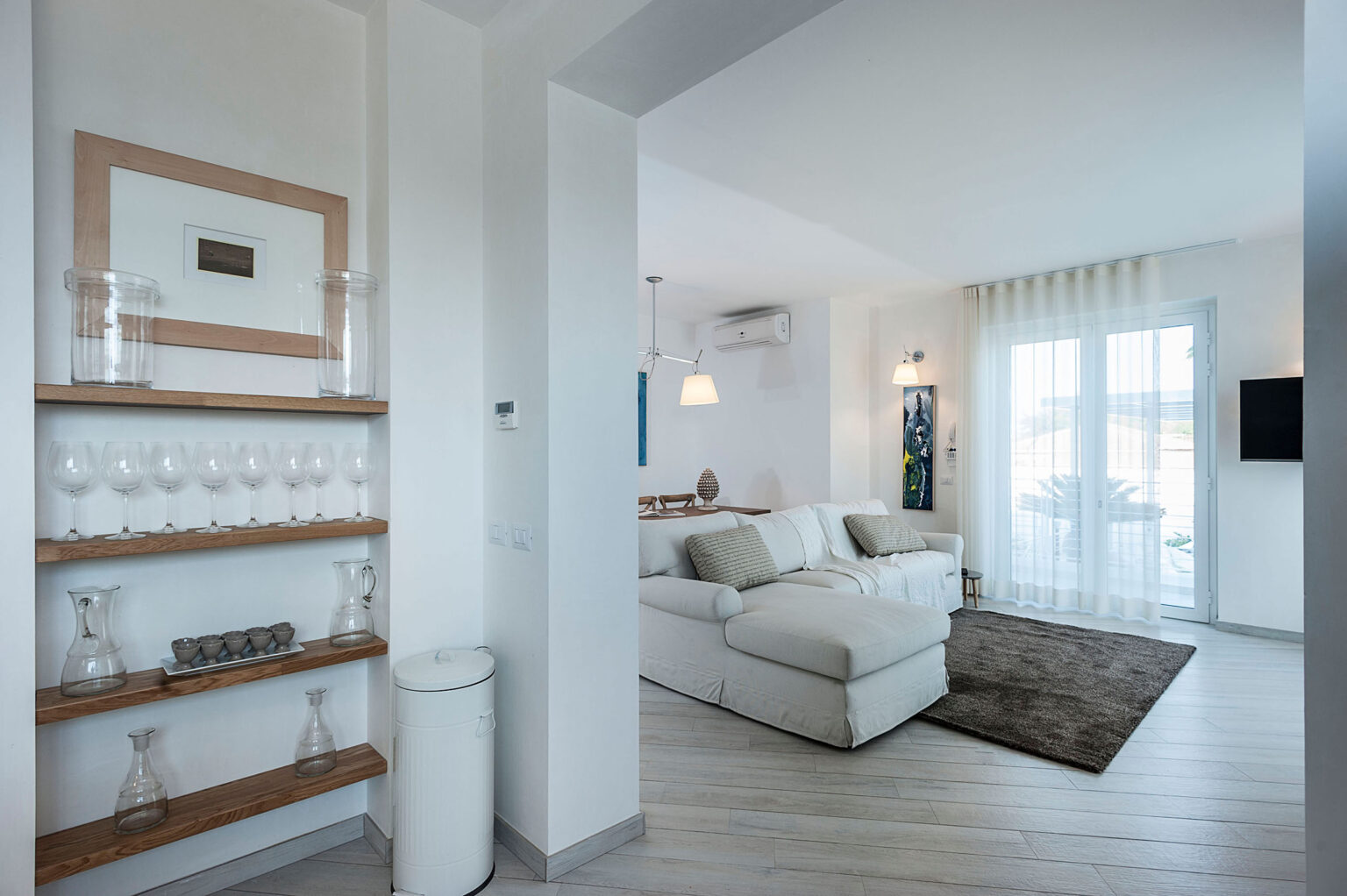 Modern living room interior featuring a cozy white sectional sofa, light wooden flooring, and large windows with sheer curtains, creating a bright and airy atmosphere. A decorative shelf with glassware adds elegance to the entryway. | Only on SopranoVillas