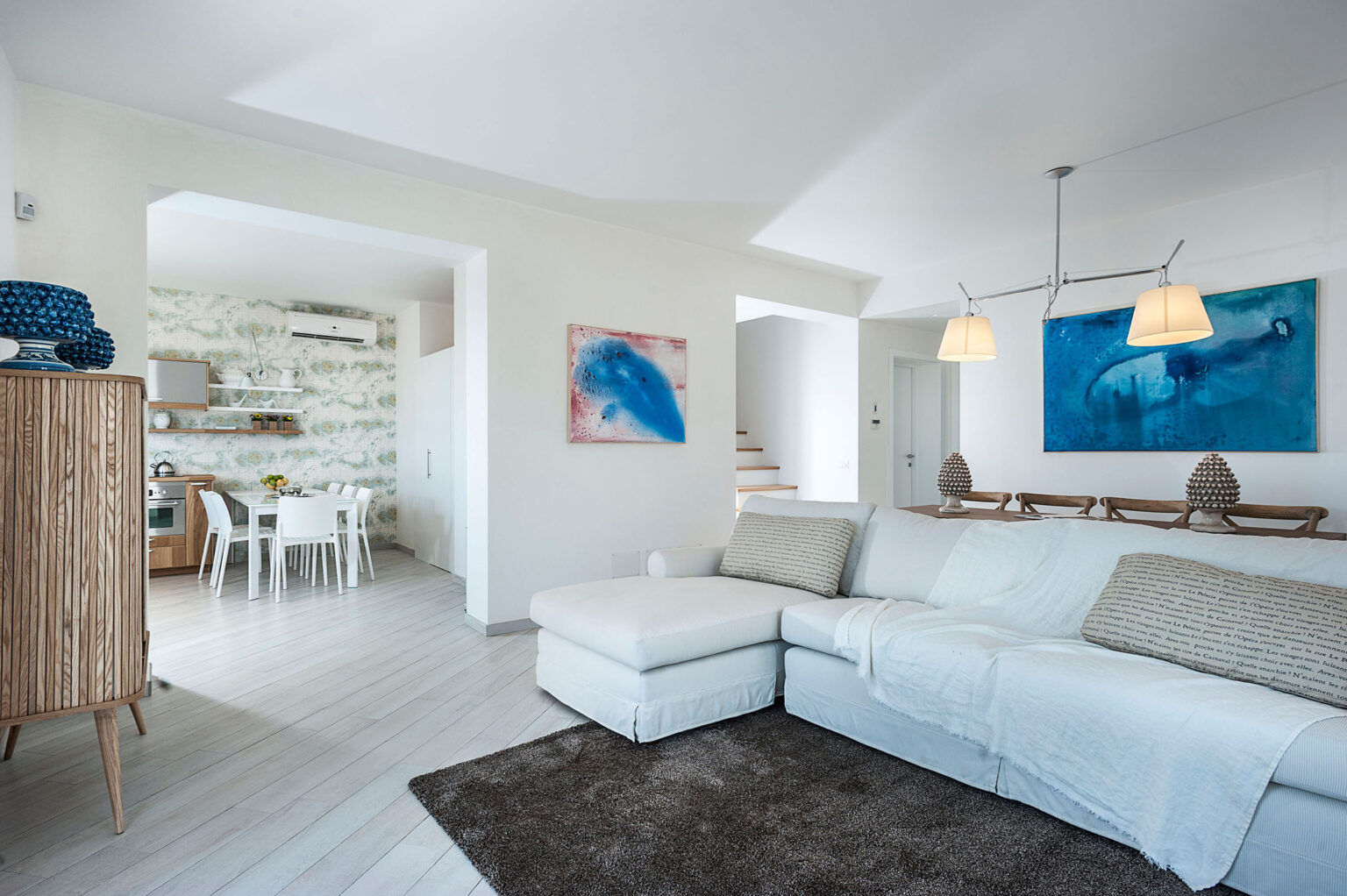 Modern living room with a white sectional sofa, wooden accents, and coastal-themed artwork. Bright, airy space with natural light and an open layout leading to a dining area. | Only on SopranoVillas