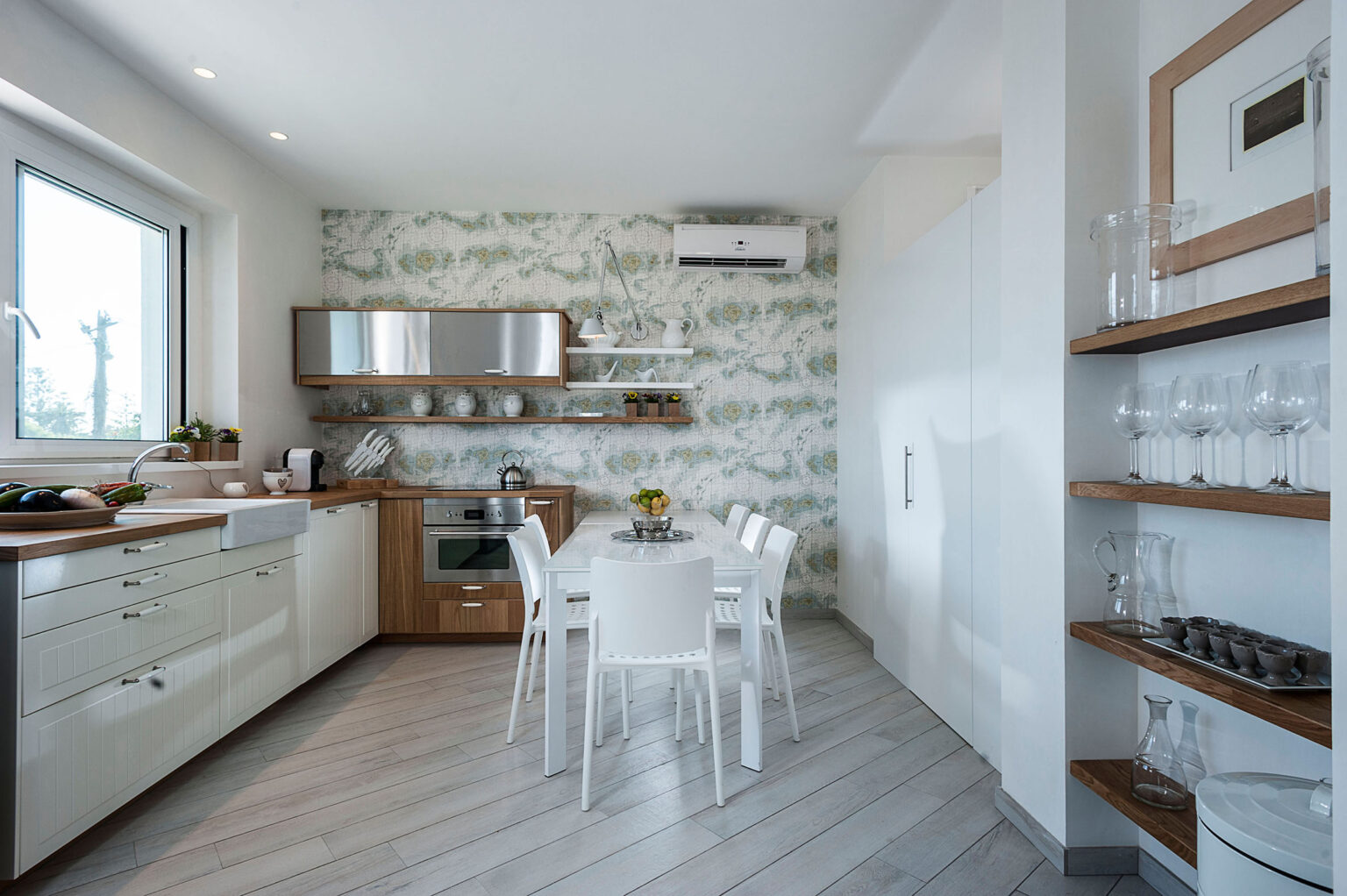 Modern kitchen featuring white cabinetry, wooden accents, and a decorative floral wallpaper. The space includes a round dining table with white chairs, stainless steel appliances, and a well-organized shelf displaying glassware. Bright and airy with natural light from a large window. | Only on SopranoVillas