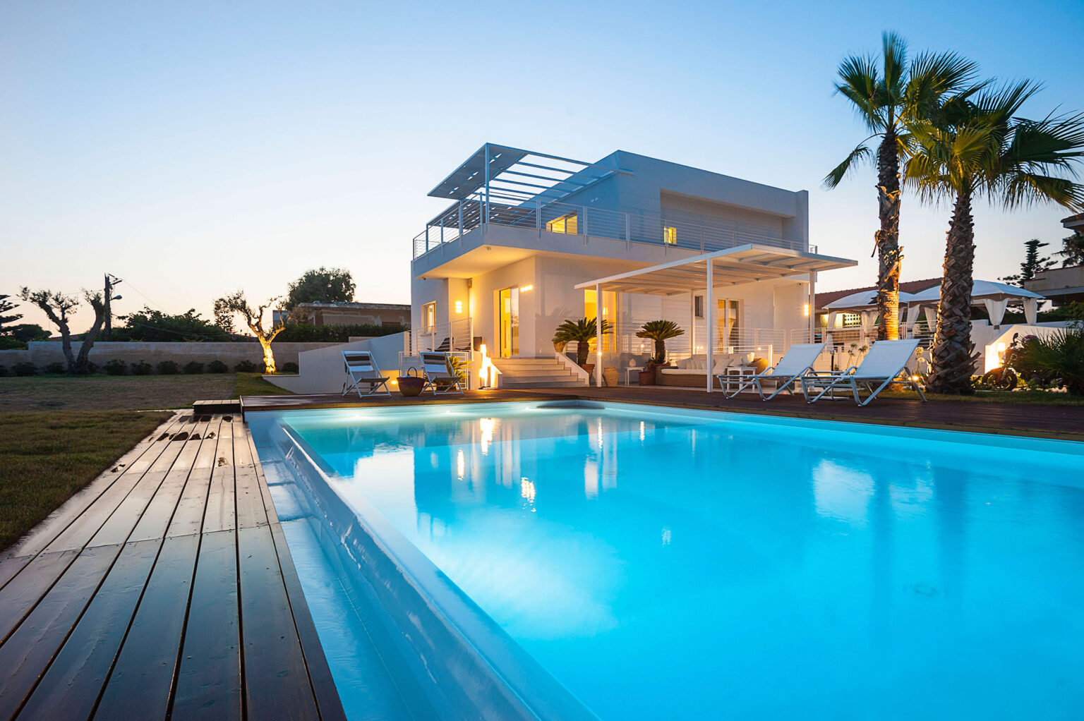 Modern villa with a swimming pool at dusk, featuring palm trees and a wooden deck, perfect for luxury outdoor living. | Only on SopranoVillas