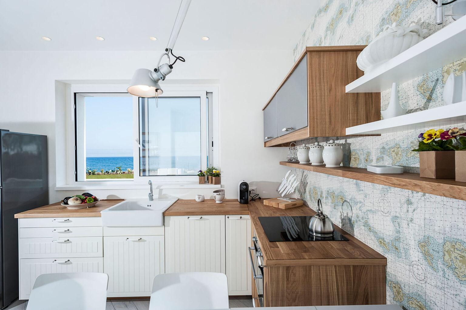 Modern kitchen with wooden cabinetry, white countertops, and a scenic ocean view through the window. Features stylish shelving with decorative items and plants, complemented by contemporary appliances. | Only on SopranoVillas