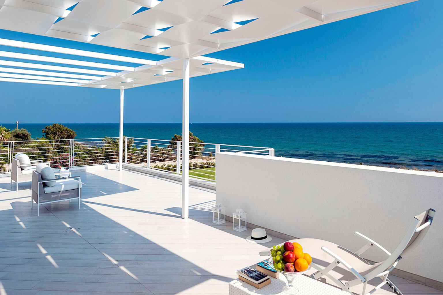Modern seaside terrace with panoramic ocean views, featuring a white pergola, lounge chairs, and a fruit bowl, perfect for relaxation and outdoor living. | Only on SopranoVillas