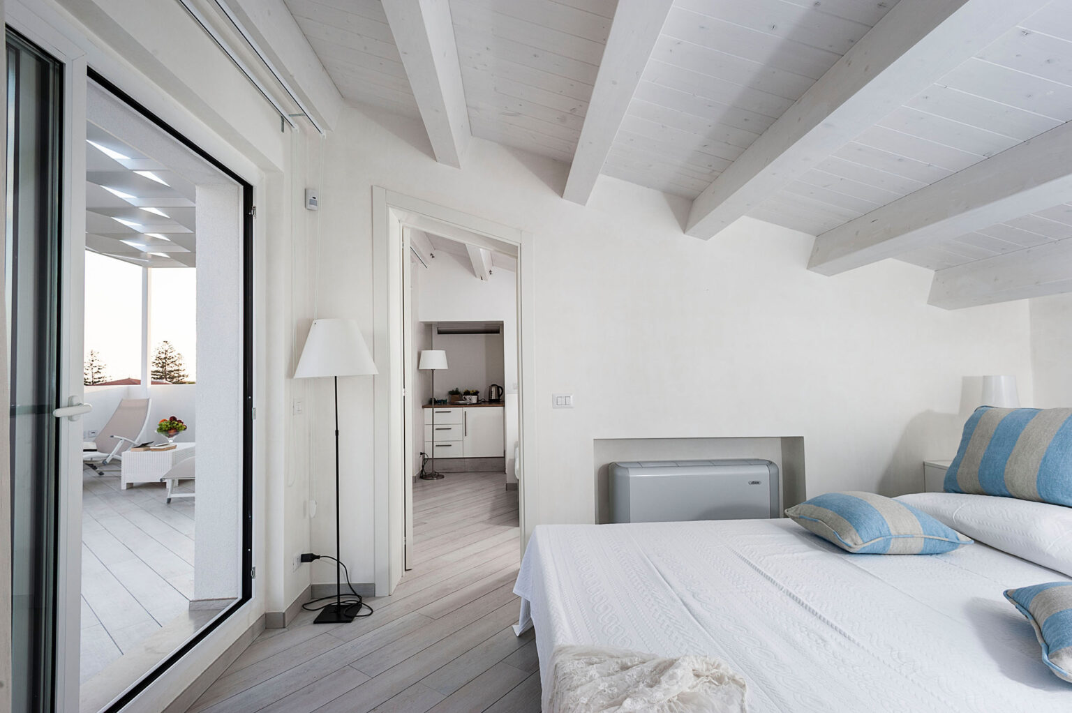 Modern bedroom interior featuring white walls and wooden beams, with a view of a bright living area through an open door. The room includes a cozy bed with blue and white striped pillows, a stylish lamp, and a sliding glass door leading to a balcony. | Only on SopranoVillas