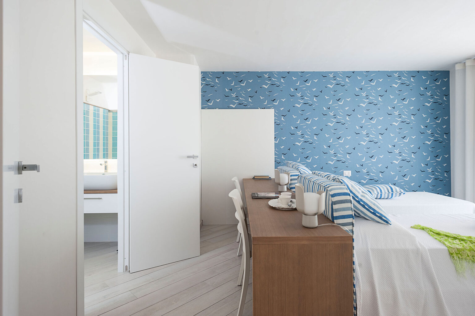 Modern hotel room featuring a blue bird-patterned accent wall, two beds with striped linens, a wooden table set for dining, and an open door leading to a bathroom with colorful tiles. The bright, airy space is designed for comfort and relaxation. | Only on SopranoVillas