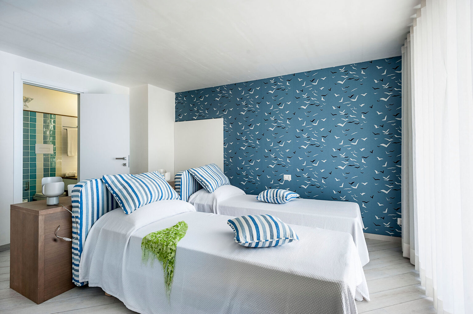 Bright and modern bedroom featuring two twin beds with blue and white striped bedding, a stylish blue patterned accent wall, and large windows allowing natural light. Ideal for a relaxing stay. | Only on SopranoVillas