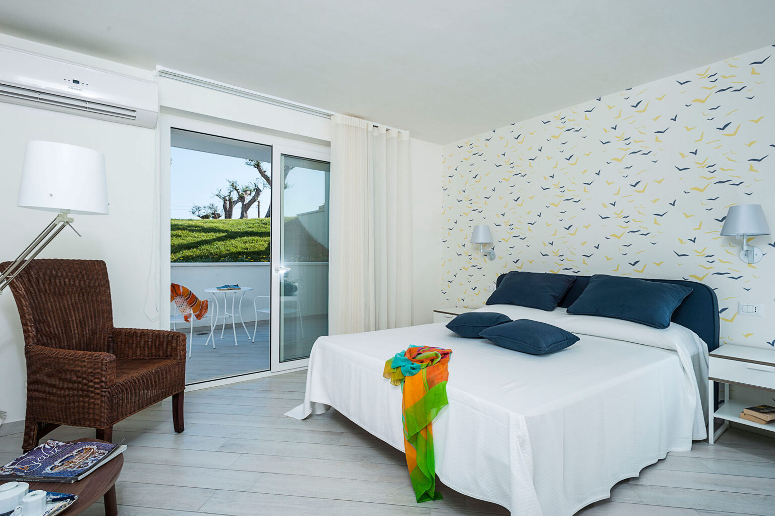 Modern hotel room featuring a queen-sized bed with blue pillows, a stylish brown chair, and a colorful throw. Large sliding doors lead to a balcony with a view of greenery, complemented by light-colored walls and wooden flooring. | Only on SopranoVillas