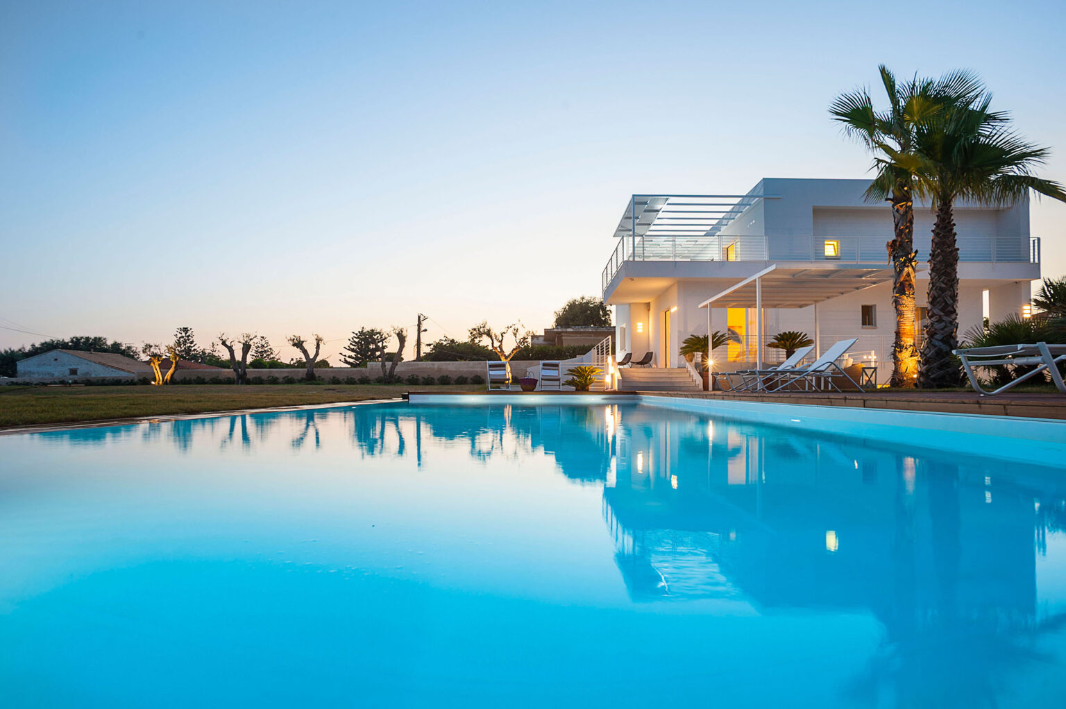 Modern villa with a swimming pool at dusk, featuring palm trees and comfortable lounge chairs, reflecting serene blue water and warm lighting. Ideal for luxury vacations or relaxation retreats. | Only on SopranoVillas