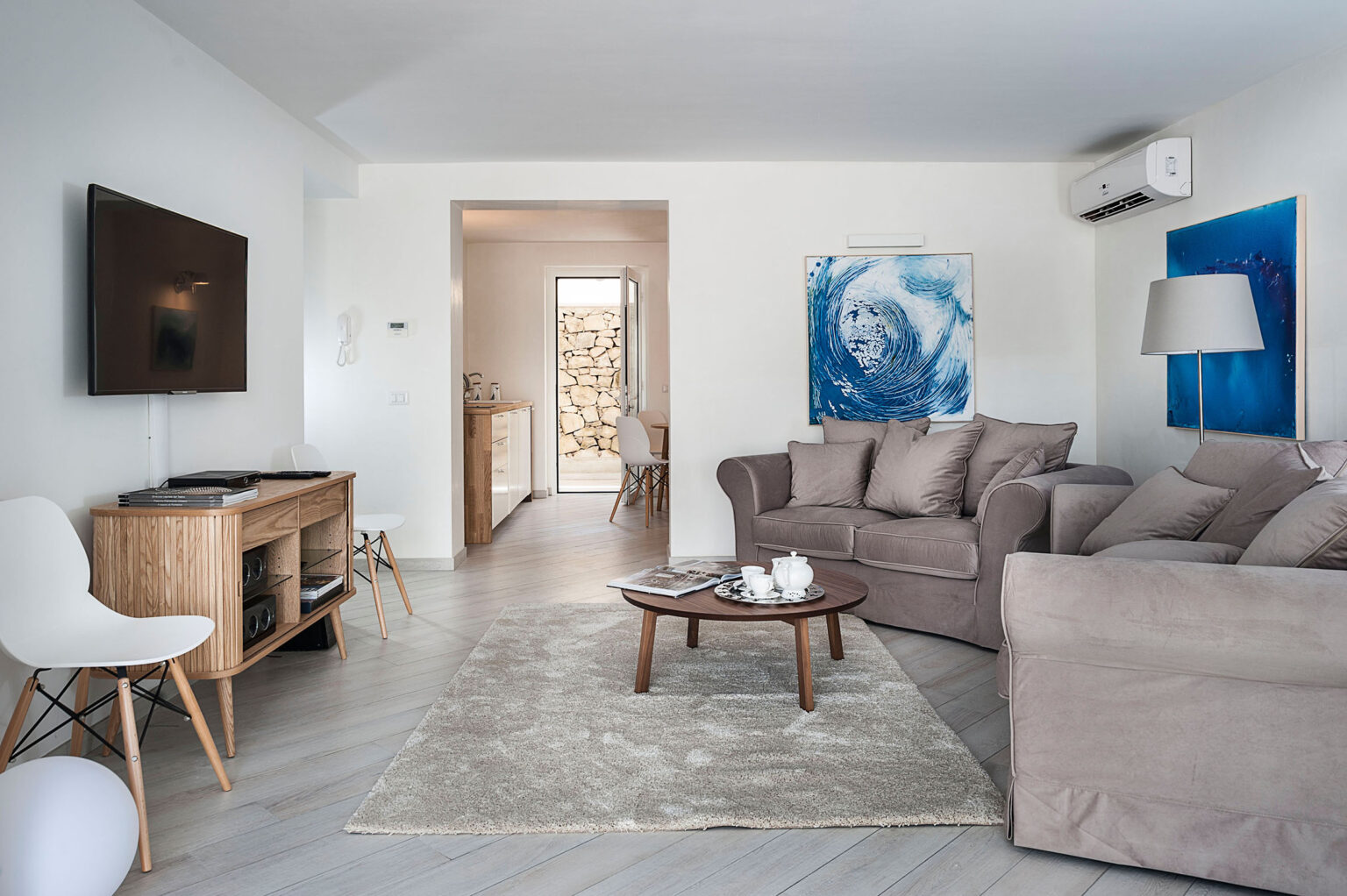 Modern living room featuring a comfortable brown sofa set, a round coffee table, and a stylish TV stand. The space is decorated with blue artwork, a neutral rug, and has ample natural light from a doorway leading to an outdoor area. | Only on SopranoVillas