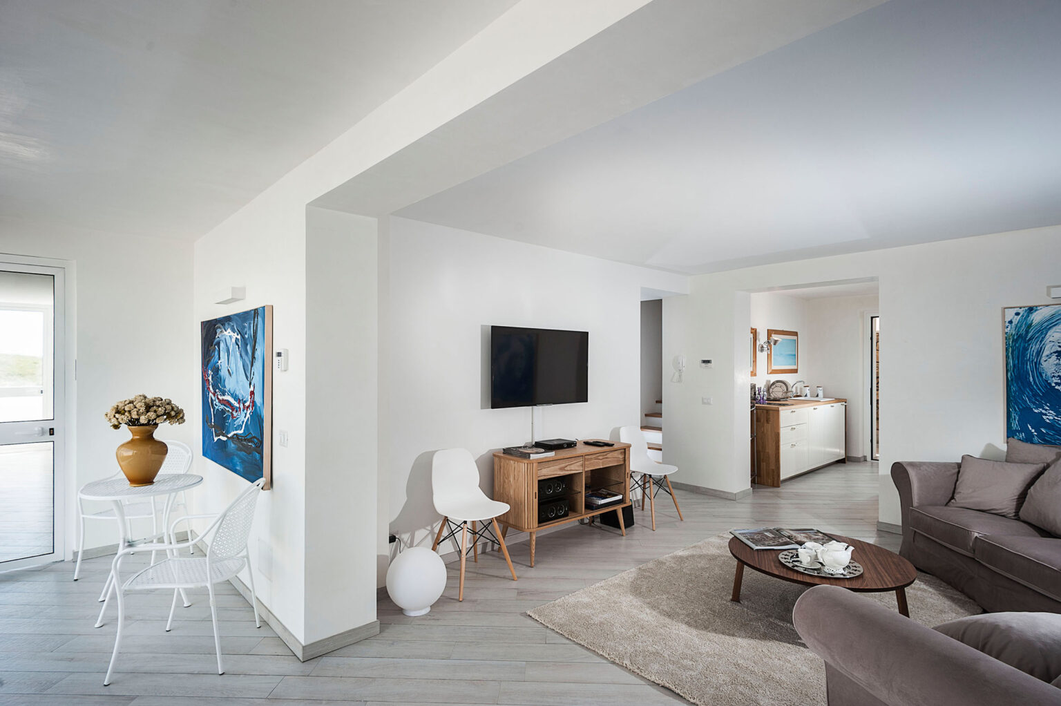 Modern living room with neutral tones, featuring a comfortable gray sofa, a wooden TV stand, and an artistic blue painting on the wall. The space includes a small dining area with a potted plant and white chairs, creating a bright and inviting atmosphere. | Only on SopranoVillas