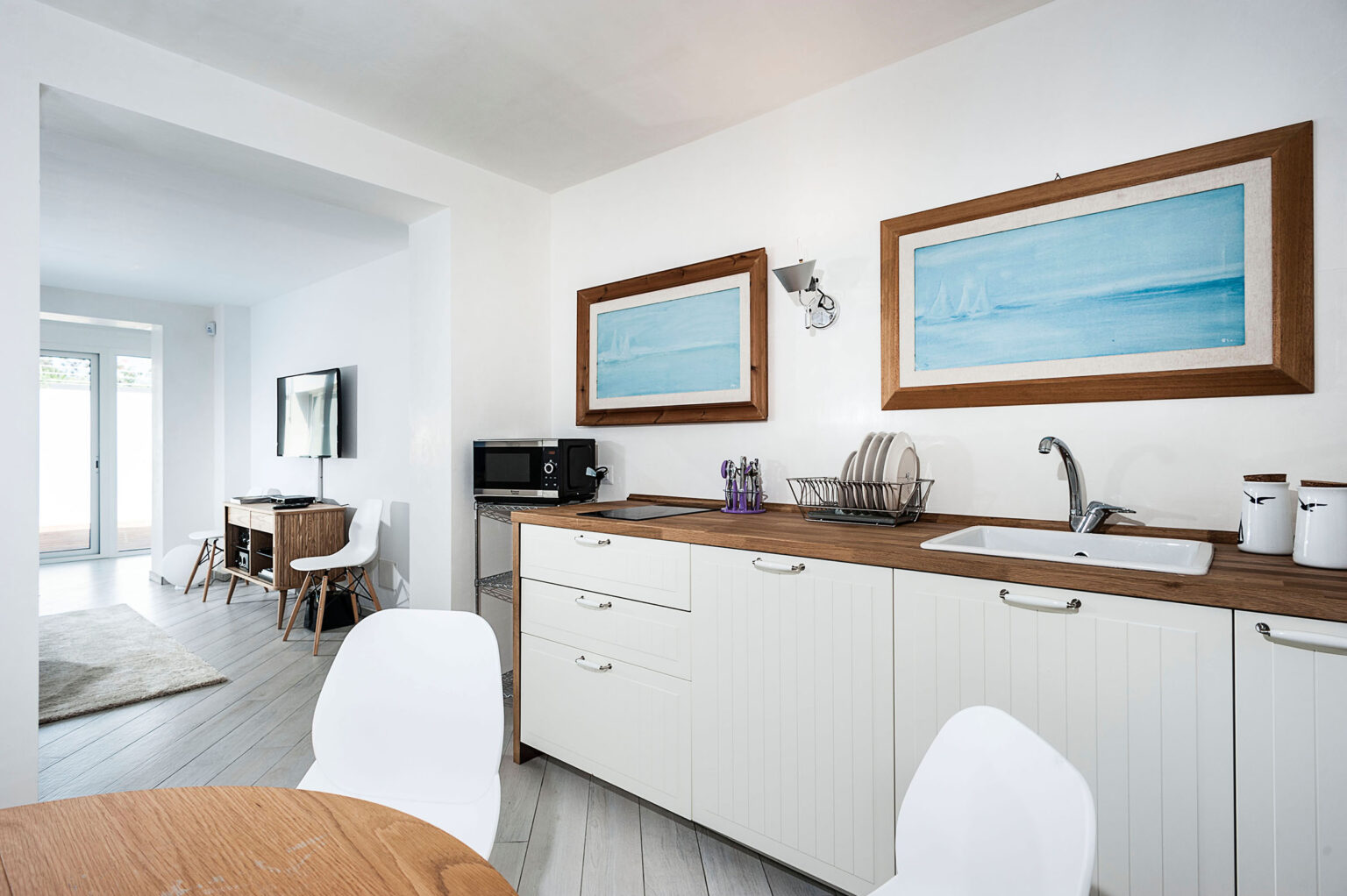 Modern kitchen interior featuring white cabinetry, wooden countertops, and blue artwork. Includes a dining area with a round table and white chairs, with natural light streaming in from nearby windows. | Only on SopranoVillas