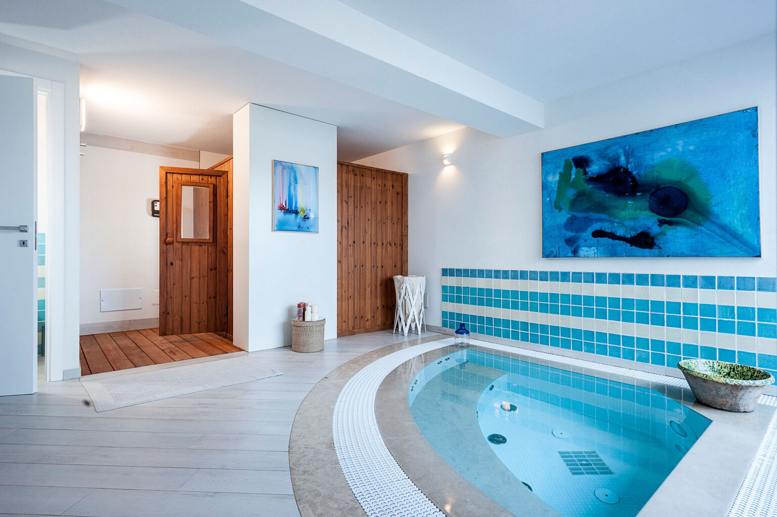 Indoor spa area featuring a circular hot tub, blue tiled accents, modern artwork on the wall, and wooden doors, creating a serene and relaxing atmosphere. | Only on SopranoVillas