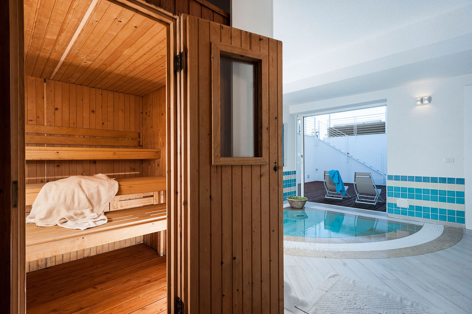 Interior view of a modern sauna with wooden benches, featuring a glass door that opens to a bright room with a small indoor pool and lounge chairs. | Only on SopranoVillas