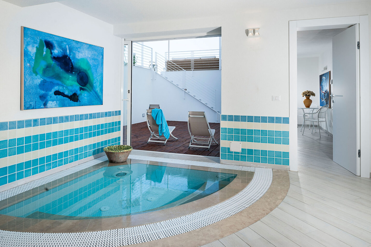 Interior view of a modern spa area featuring a circular indoor jacuzzi surrounded by blue and white tiles, leading to an outdoor deck with lounge chairs. A vibrant abstract painting adorns the wall, enhancing the serene atmosphere. Natural light floods the space through large glass doors. | Only on SopranoVillas
