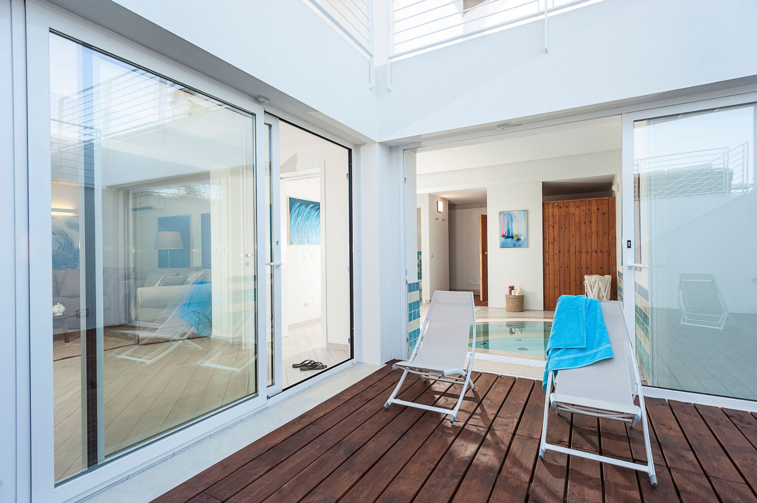 Modern outdoor patio area featuring two lounge chairs with a blue towel, overlooking a stylish interior with large glass doors, a cozy living space, and a small pool. Ideal for relaxation and enjoying a tranquil atmosphere. | Only on SopranoVillas