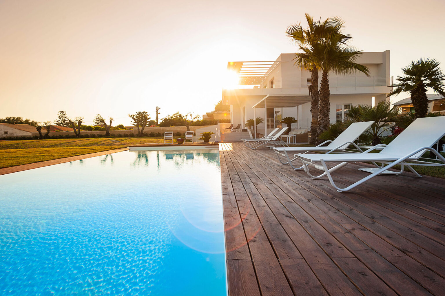 Modern villa with a swimming pool at sunset, featuring wooden decking and lounge chairs surrounded by palm trees and landscaped gardens. Ideal for luxury vacation rentals or outdoor relaxation. | Only on SopranoVillas