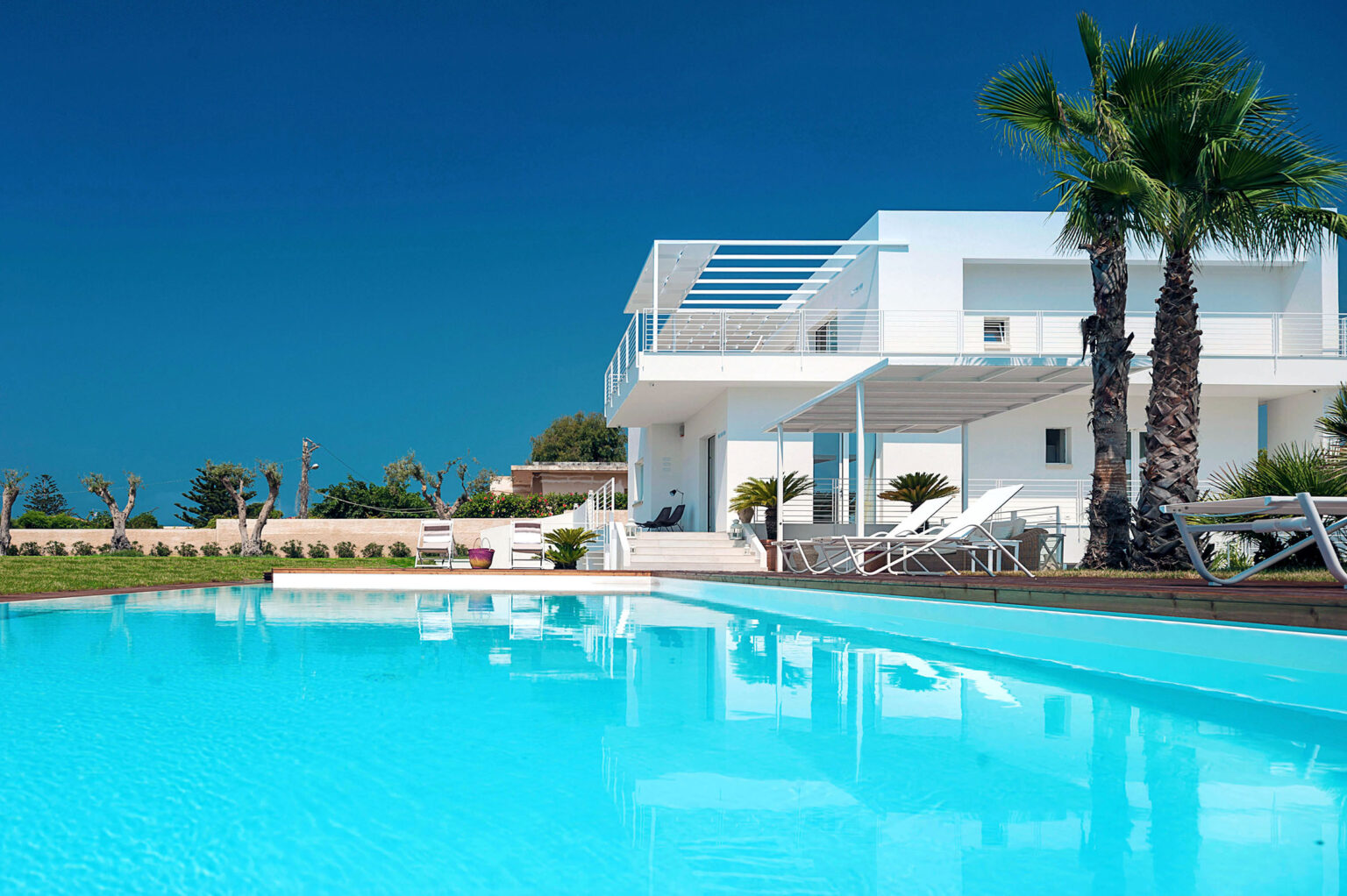 Modern white villa with a swimming pool, surrounded by palm trees and lush greenery, under a clear blue sky. The pool reflects the villa, creating a serene outdoor space ideal for relaxation and leisure. | Only on SopranoVillas