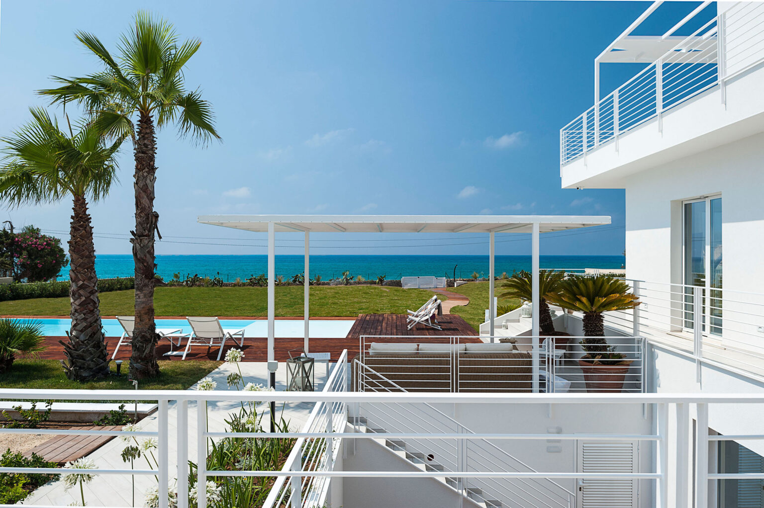 Modern coastal villa with a spacious terrace, palm trees, and a swimming pool overlooking the ocean under a clear blue sky. | Only on SopranoVillas