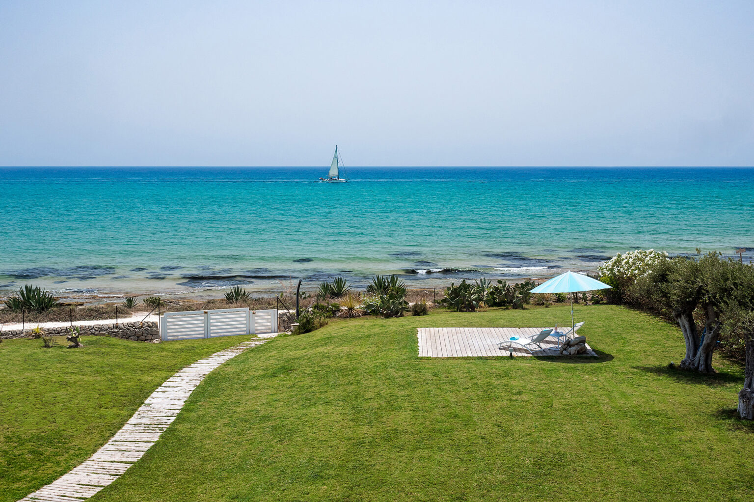 Scenic view of a coastal landscape featuring a lush green lawn, a sailboat on the turquoise sea, and a shaded patio area with an umbrella. Perfect for vacation rentals or beach getaways. | Only on SopranoVillas