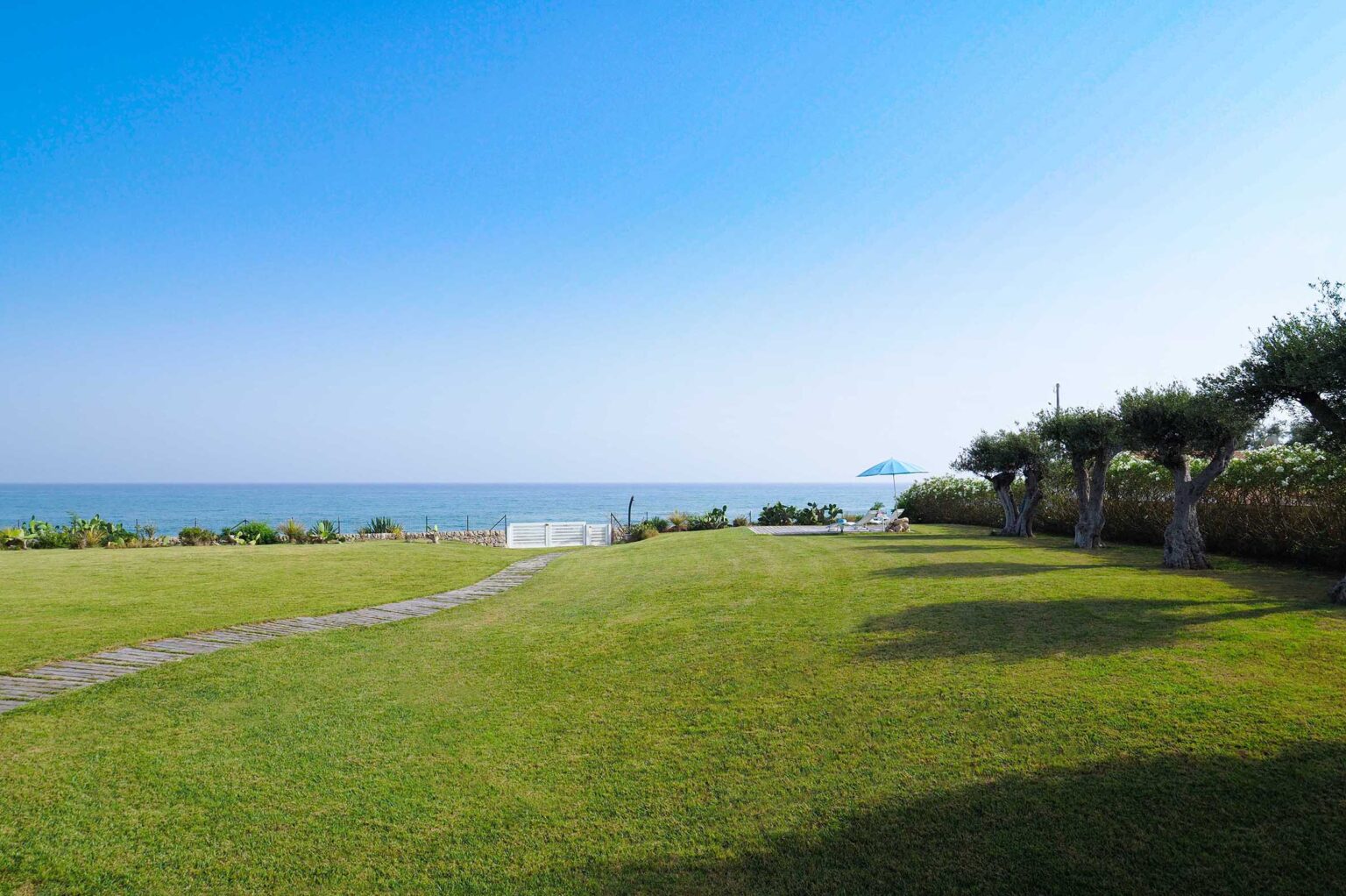 Lush green lawn leading to a serene beach with a clear blue sky, featuring a pathway and a sun umbrella near lounge chairs by the water. Ideal for relaxation and outdoor activities. | Only on SopranoVillas