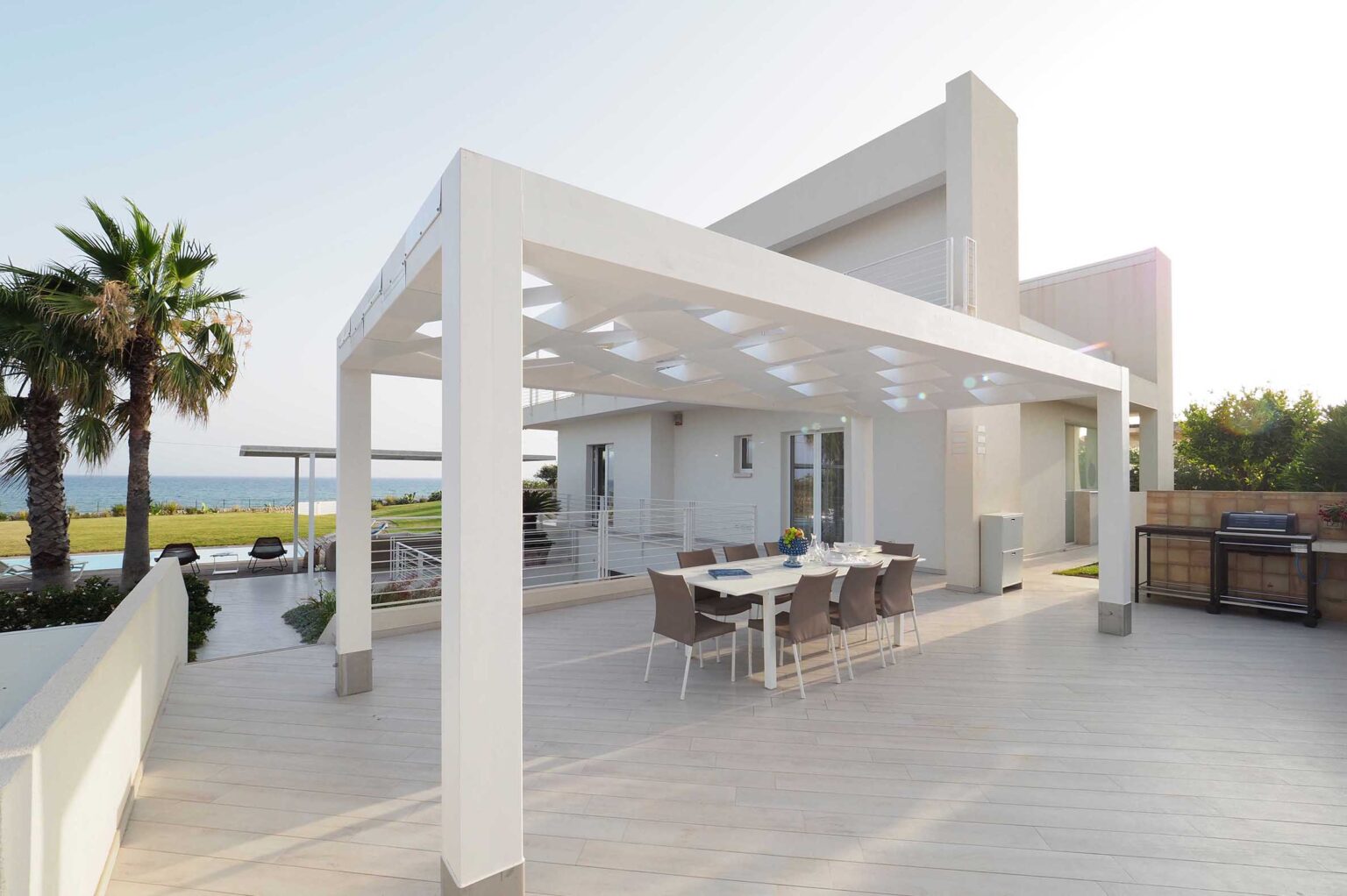 Modern outdoor patio with a white pergola, dining table, and chairs overlooking a scenic view of the ocean and palm trees. Ideal for entertaining and relaxation in a luxurious setting. | Only on SopranoVillas