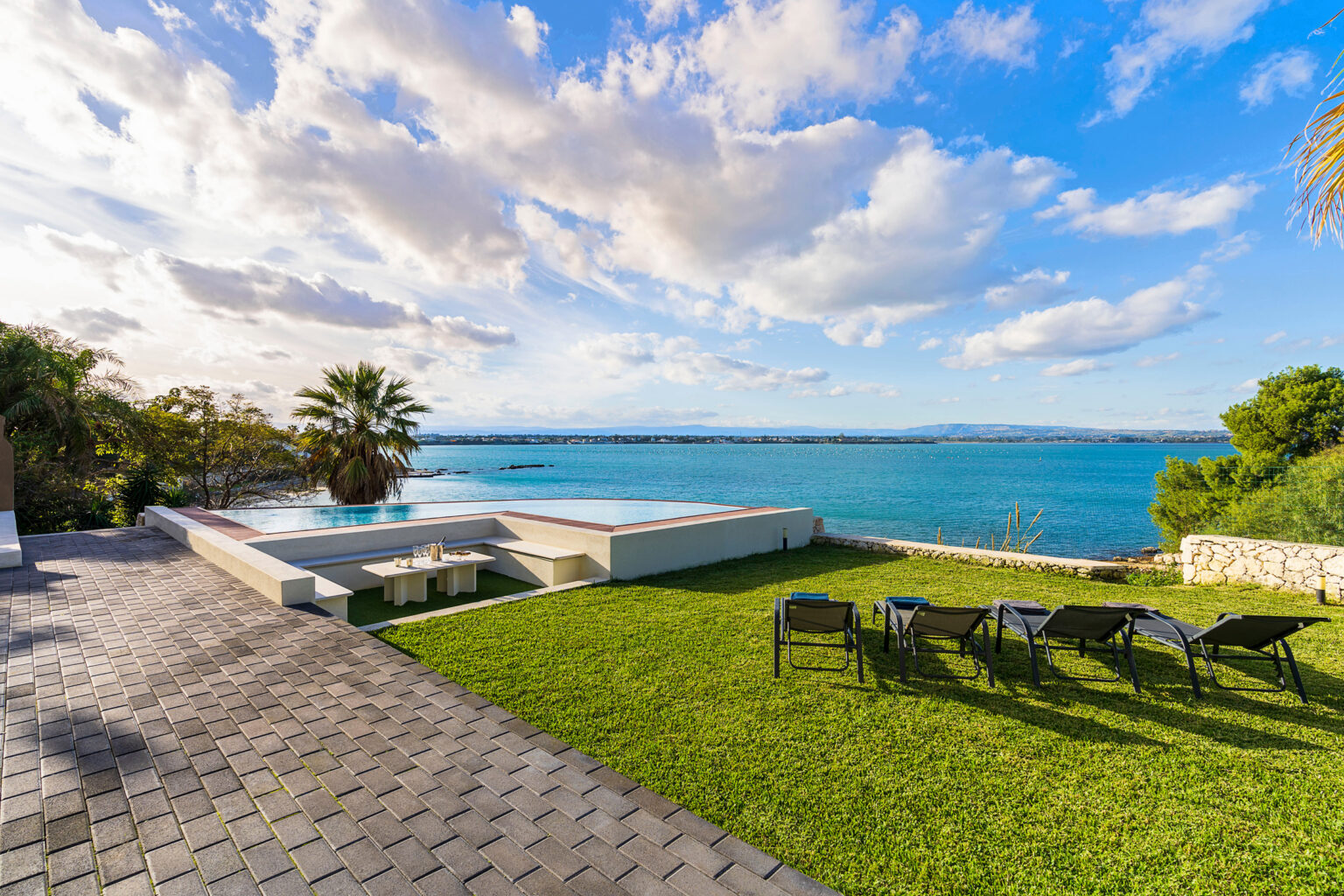 Scenic view of a coastal property featuring a well-manicured lawn, lounge chairs, and a swimming pool overlooking the ocean, with palm trees and a clear blue sky in the background. | Only on SopranoVillas