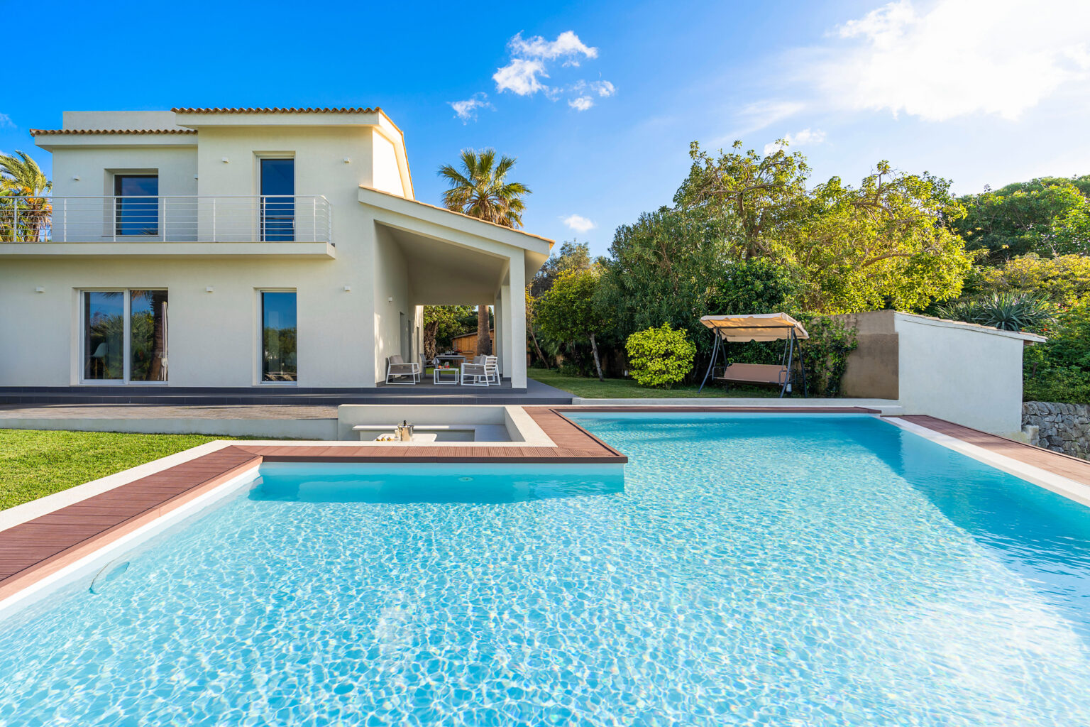 Modern villa with a swimming pool, surrounded by lush greenery and palm trees, featuring a spacious patio and outdoor seating area under a shaded porch. Bright blue sky enhances the serene atmosphere. | Only on SopranoVillas
