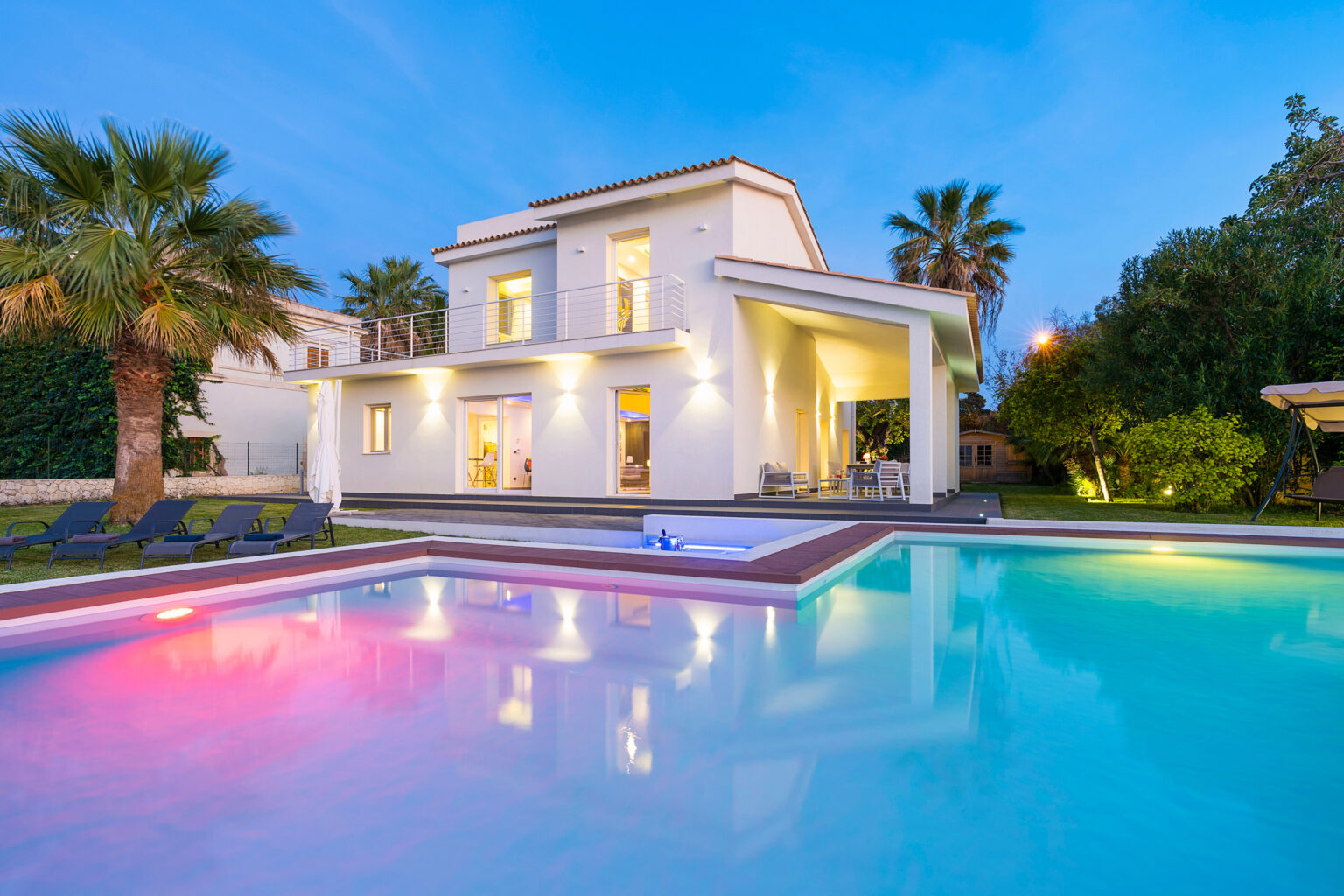 Modern villa with a swimming pool, illuminated at dusk, surrounded by palm trees and lush greenery. Ideal for luxury living and relaxation. | Only on SopranoVillas
