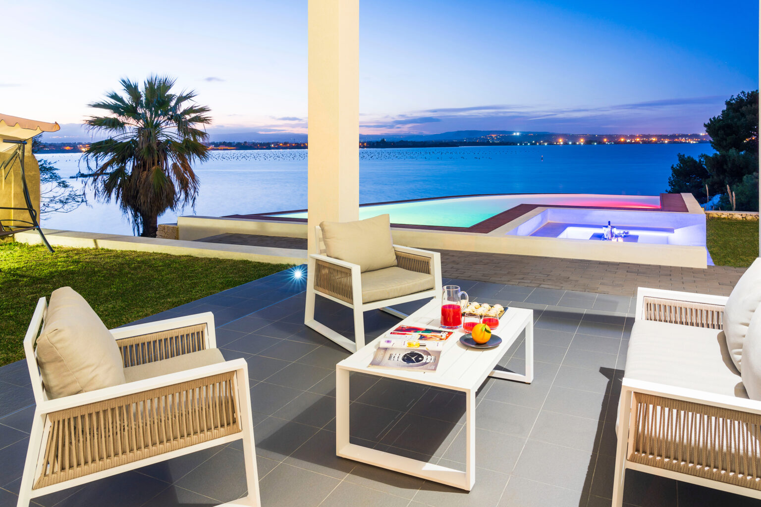 Modern outdoor living space featuring comfortable seating, a coffee table with snacks, and a private hot tub overlooking a serene lake at sunset. Ideal for relaxation and entertainment. | Only on SopranoVillas