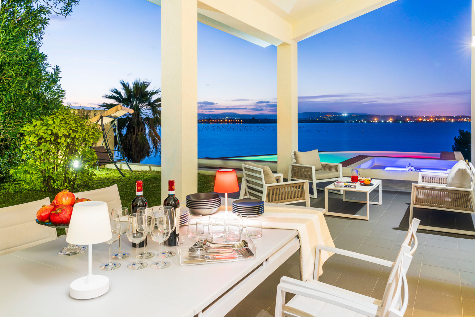 Luxurious outdoor terrace with a dining table featuring apples, wine bottles, and glassware, overlooking a serene waterfront at dusk, with comfortable seating and a hot tub illuminated by colorful lights. | Only on SopranoVillas