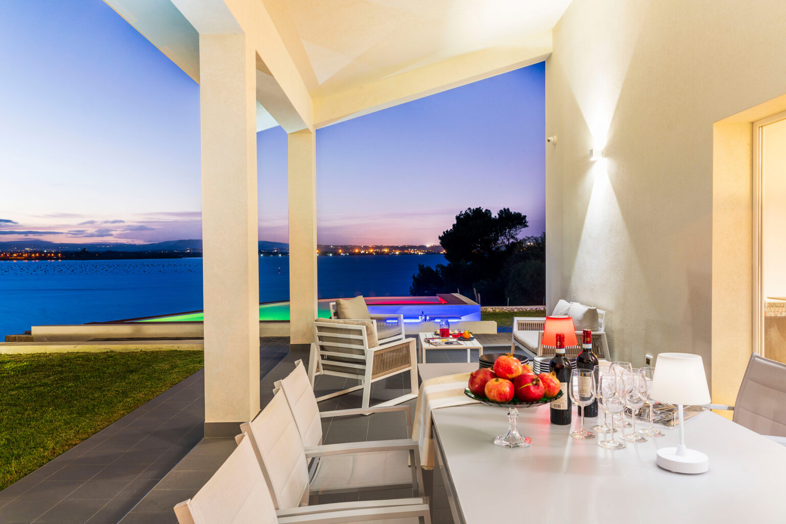 Elegant outdoor dining area with a stunning sunset view over the water, featuring a table set with fresh fruit and wine, surrounded by modern seating and a relaxing atmosphere. | Only on SopranoVillas