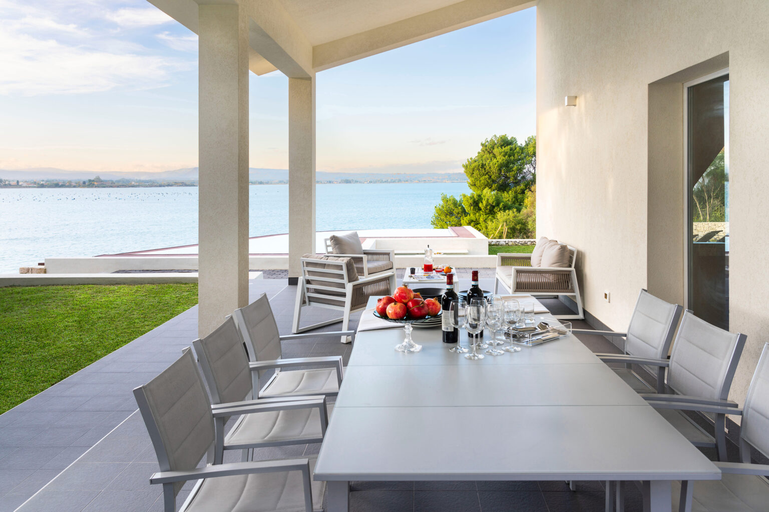 Modern outdoor dining area with a table set for a meal, featuring wine glasses and fresh fruit, overlooking a scenic view of the water and greenery. Ideal for entertaining and enjoying nature. | Only on SopranoVillas