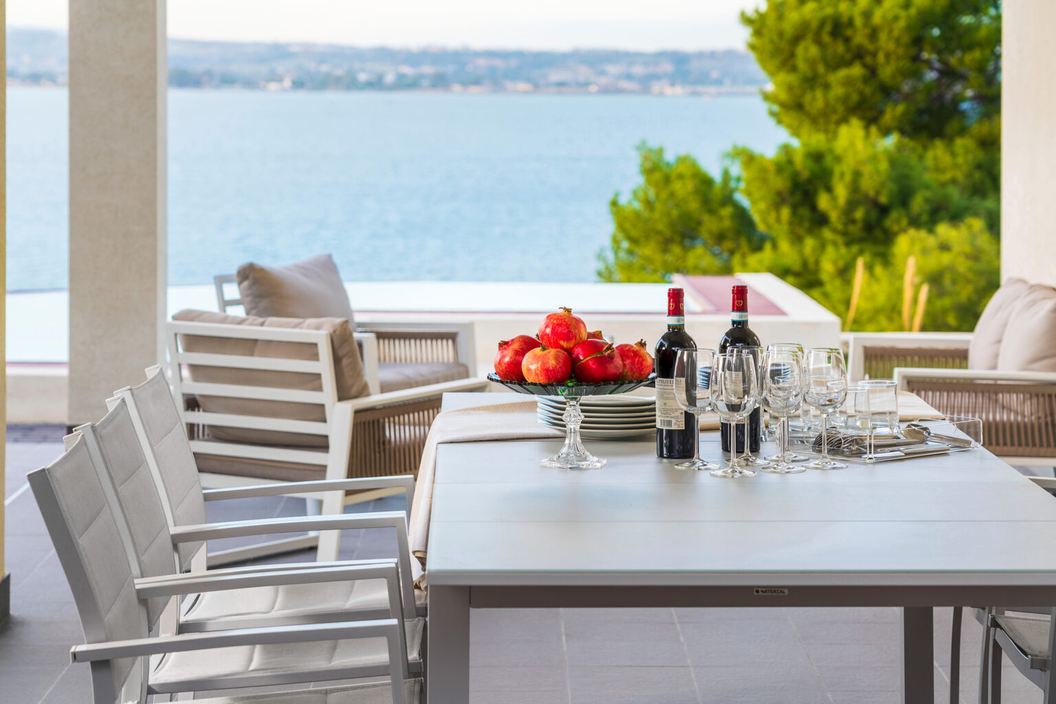 Outdoor dining area with a table set for a meal, featuring wine bottles and glasses, overlooking a scenic waterfront view and lush greenery. Perfect for entertaining guests in a relaxing atmosphere. | Only on SopranoVillas
